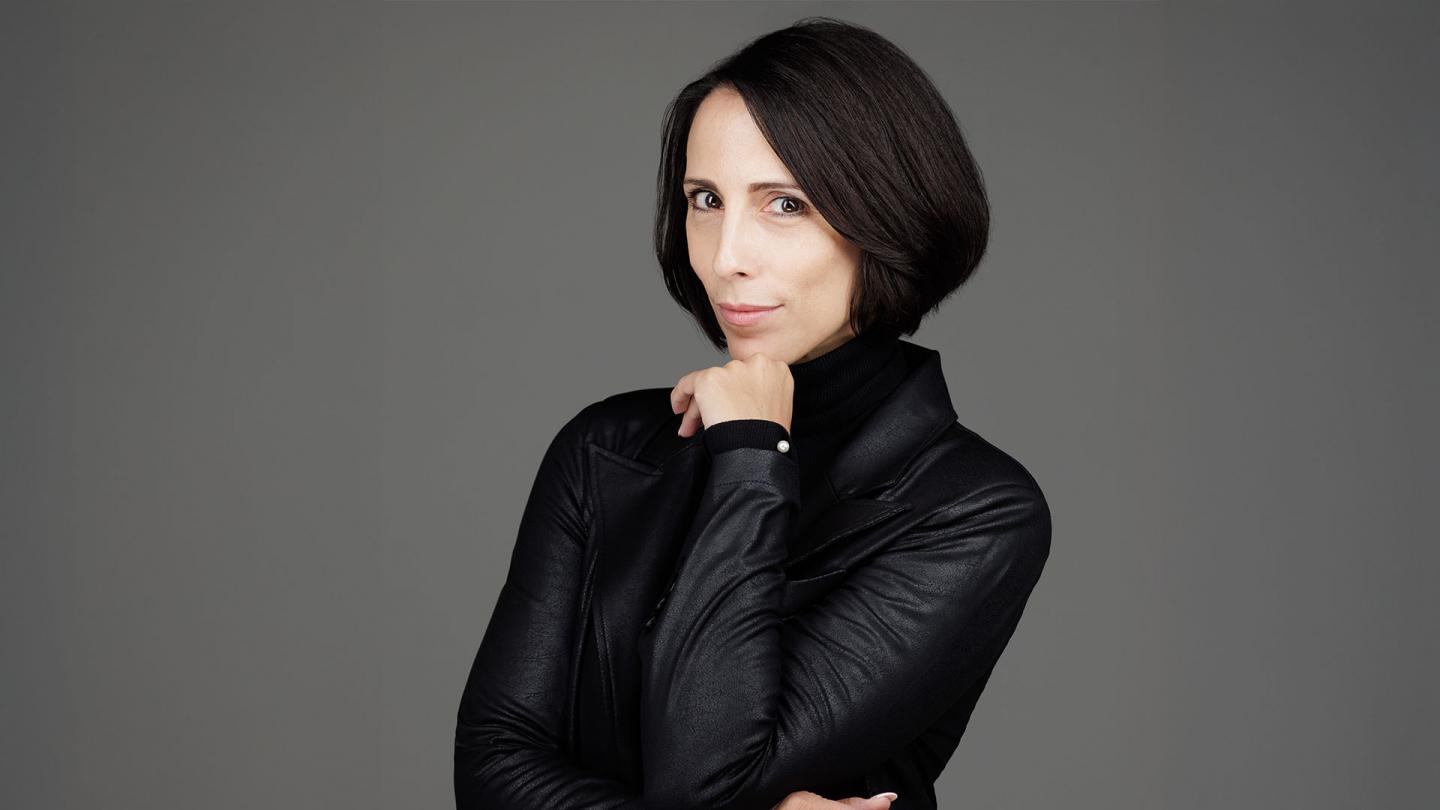 Woman with dark hair, a wan smile, black jacket and her arms crossed with one hand under her chin