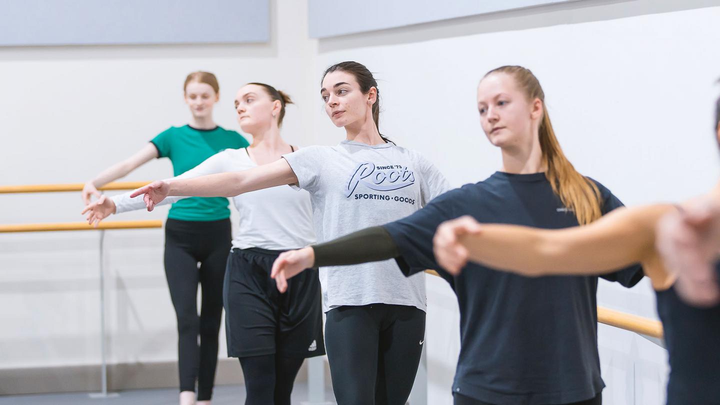 Student dancers at the barre, holding on with one hand, their other arm outstretched