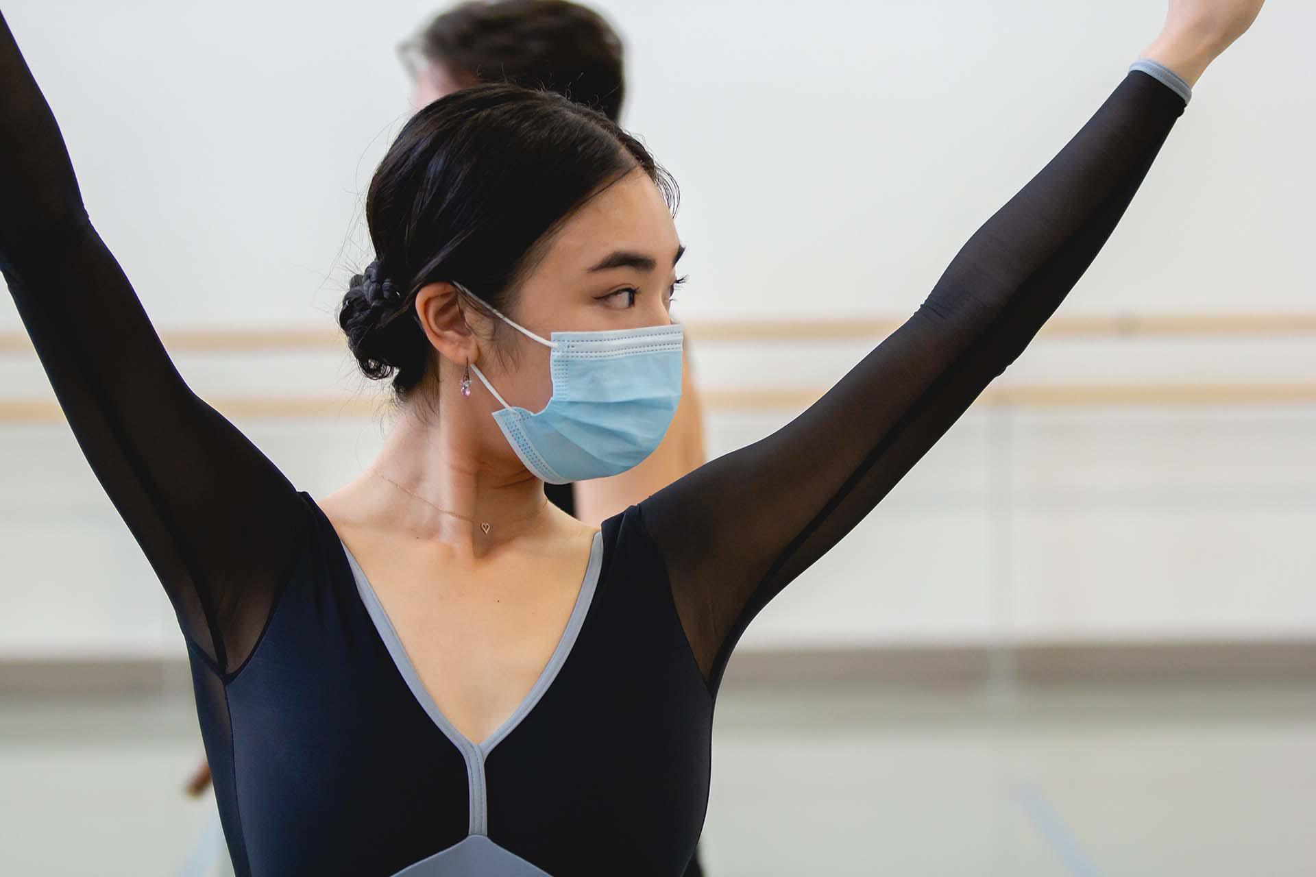 A female dancer in a face mask looks to her left while holding her hands above her head