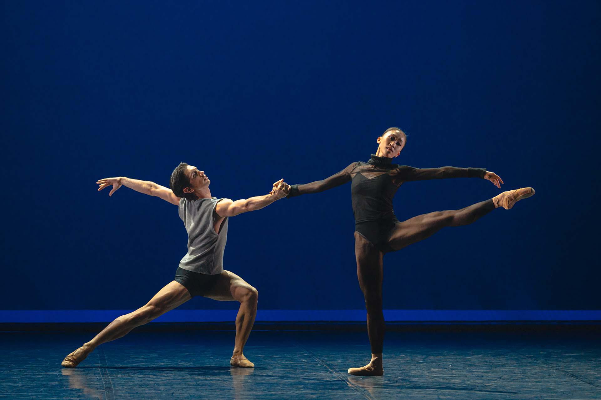 Male dancer holding the hand of a female dancer as he stretches a leg back and she has one leg outstretched