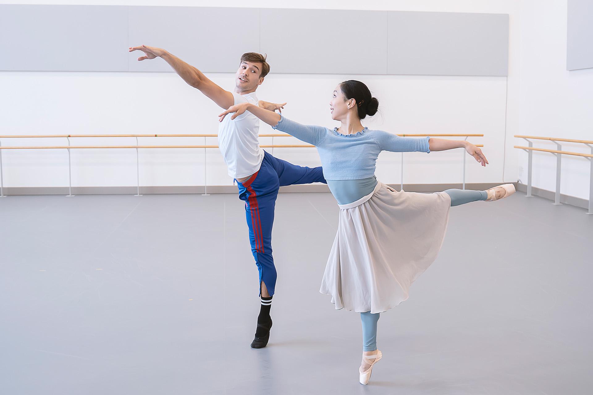 Two dancers mirror each other's pose, on one foot, leg stretched behind them, arm stretched in front of them, looking at one another