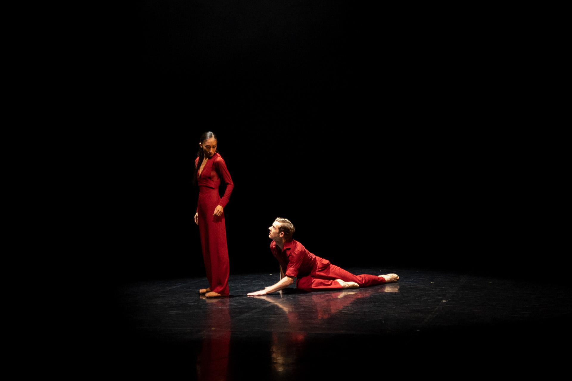 Two dancers dress in red, one is on the floor looking up at the other who is turned away but sees him over her shoulder
