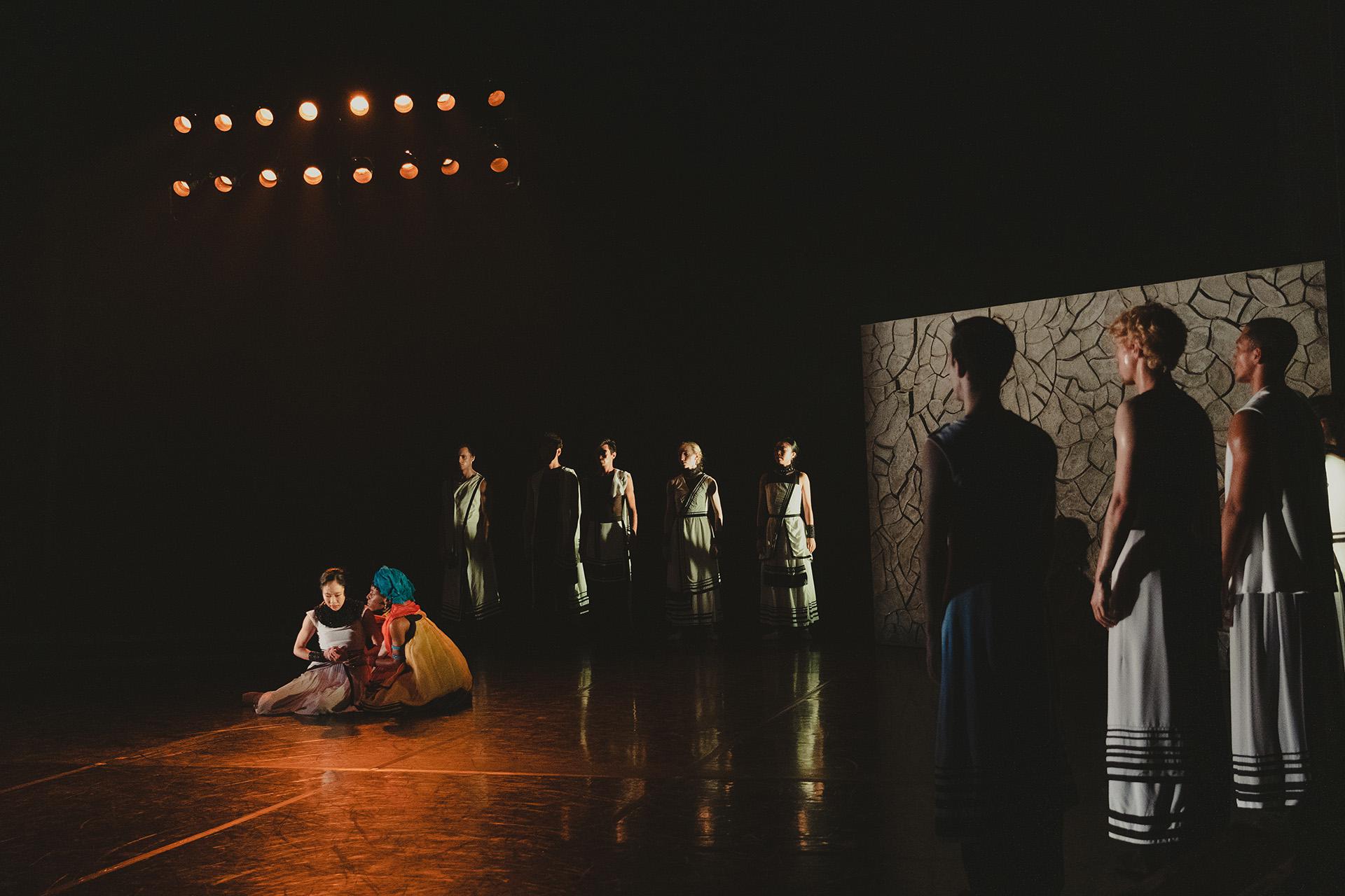 Two people sit on the stage comforting each other, others look on with concern