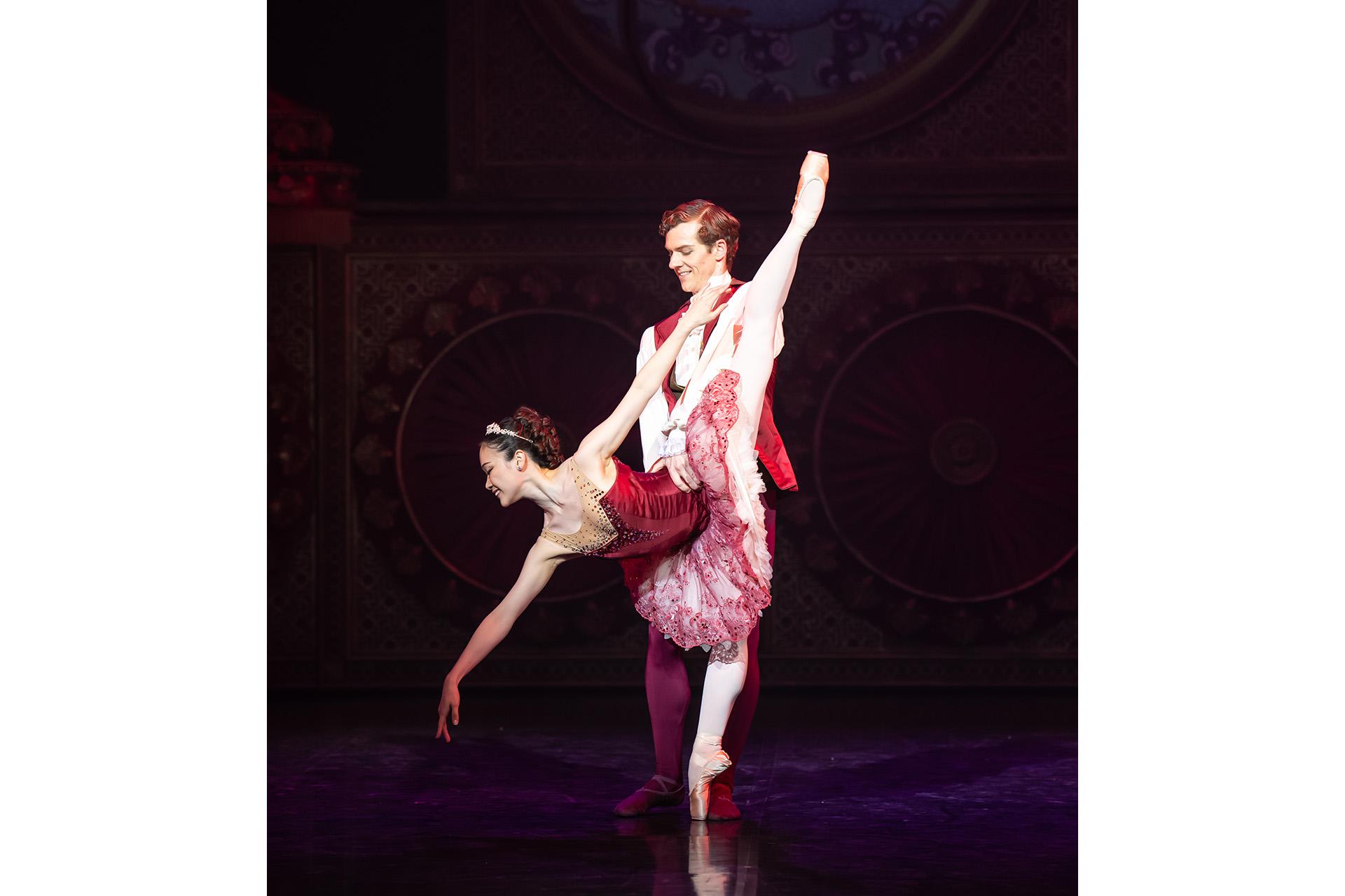 A dancer supports another as she extends one of her legs behind her while reaching to the floor