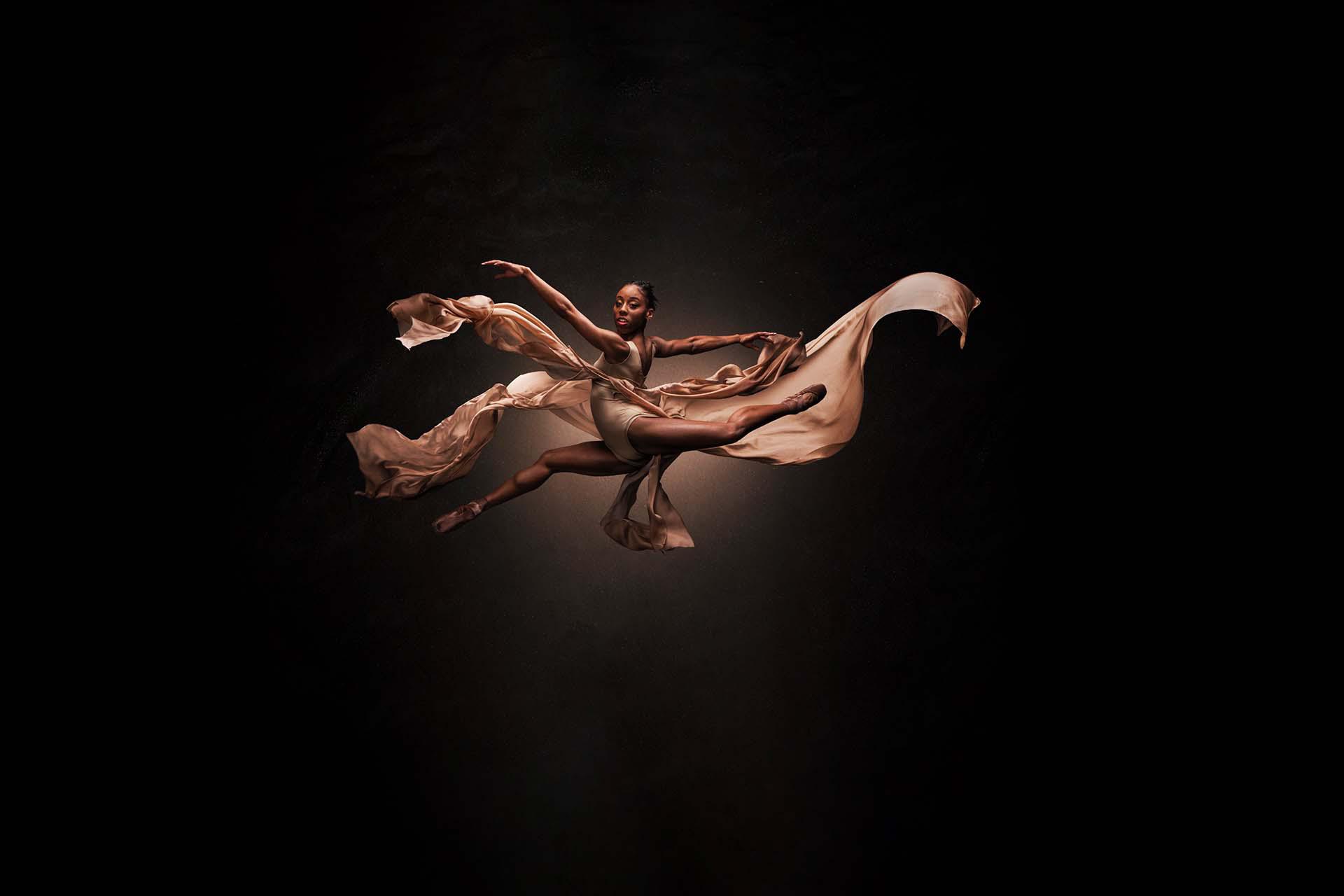 Dancer leaping across the frame while looking directly at the viewer, wearing peach and with fabrics flowing in the wind