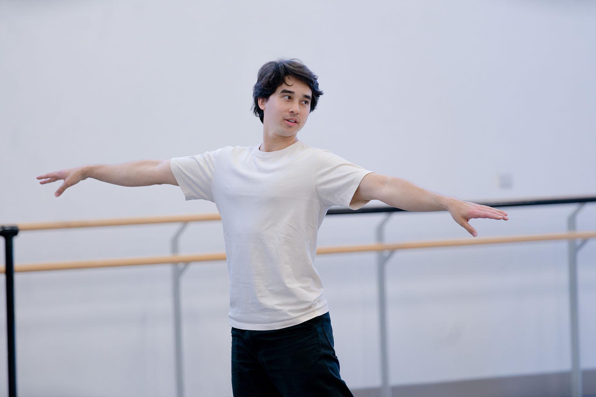 Benjamin Ella in a white T-shirt and black trousers with his arms straight out either side of this body and looking to his left