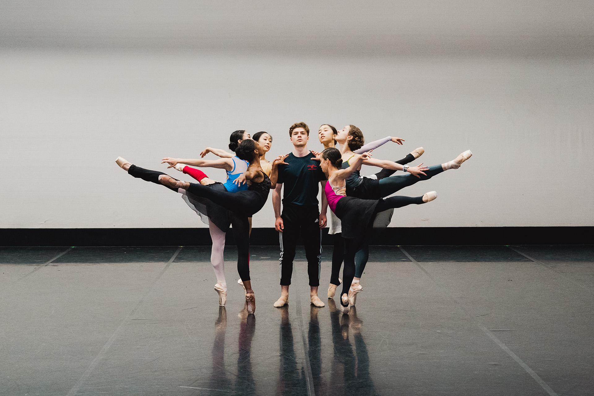 Male dancer stands emotionless while surrounded by six dancers, all arabesque and en pointe, supporting themselves by placing their free hand on his shoulder
