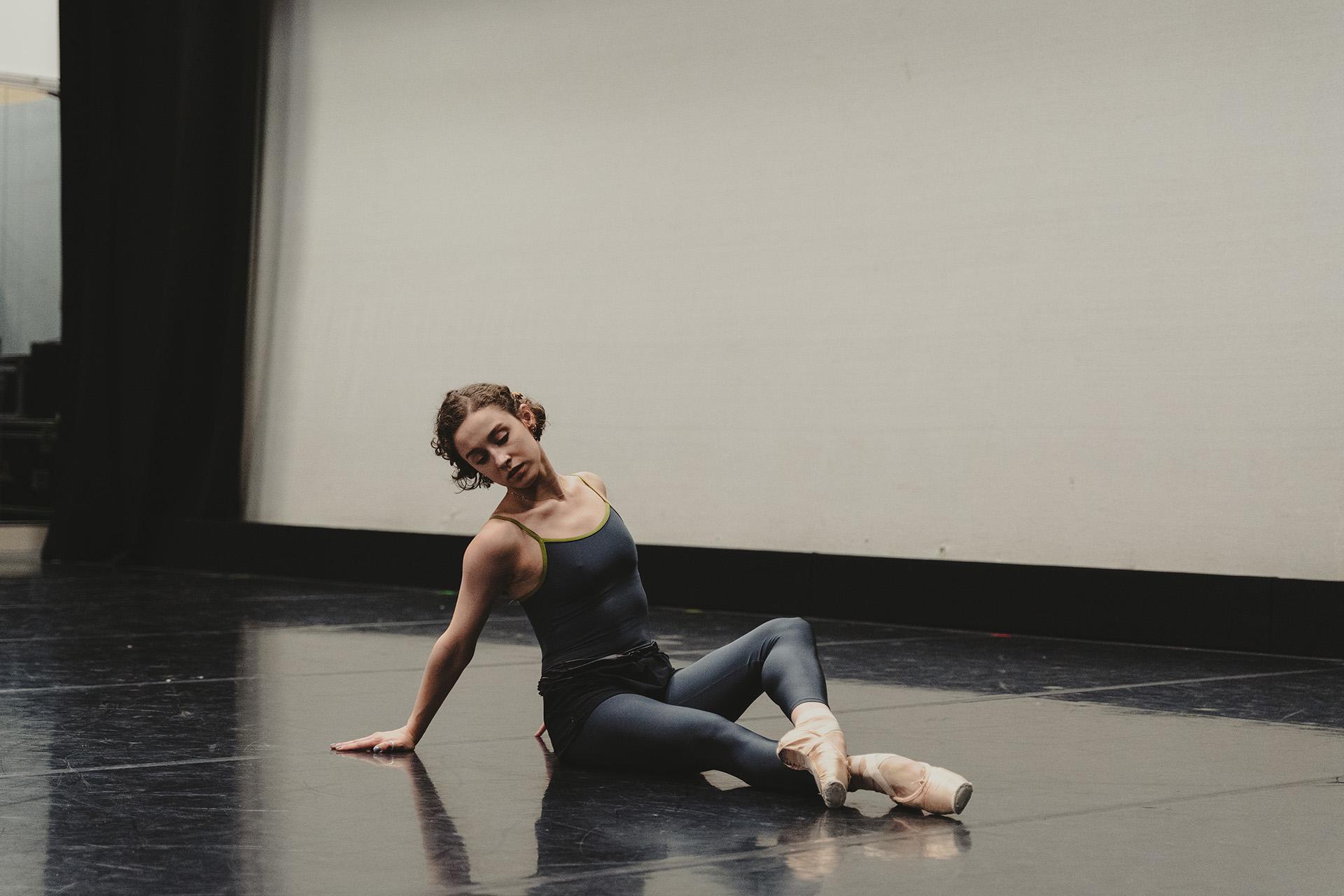 Dancer wearing grey and pale pink pointe shoes sits on the floor in a deliberate pose, torso upright but leaning back on her hands