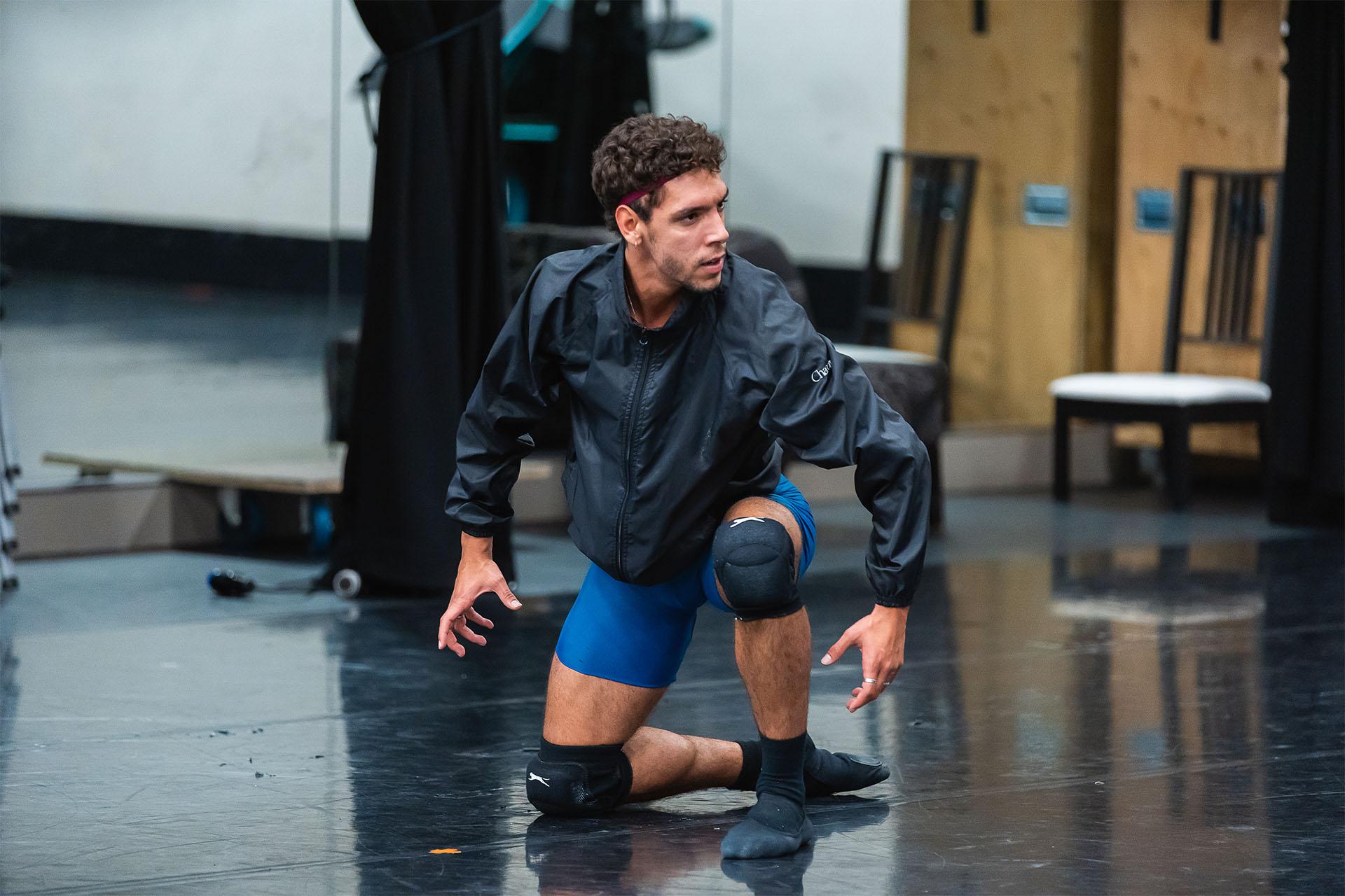 Male dancer with a dark top and blue shorts menacingly kneels on the dance floor as he rehearses as the Beast