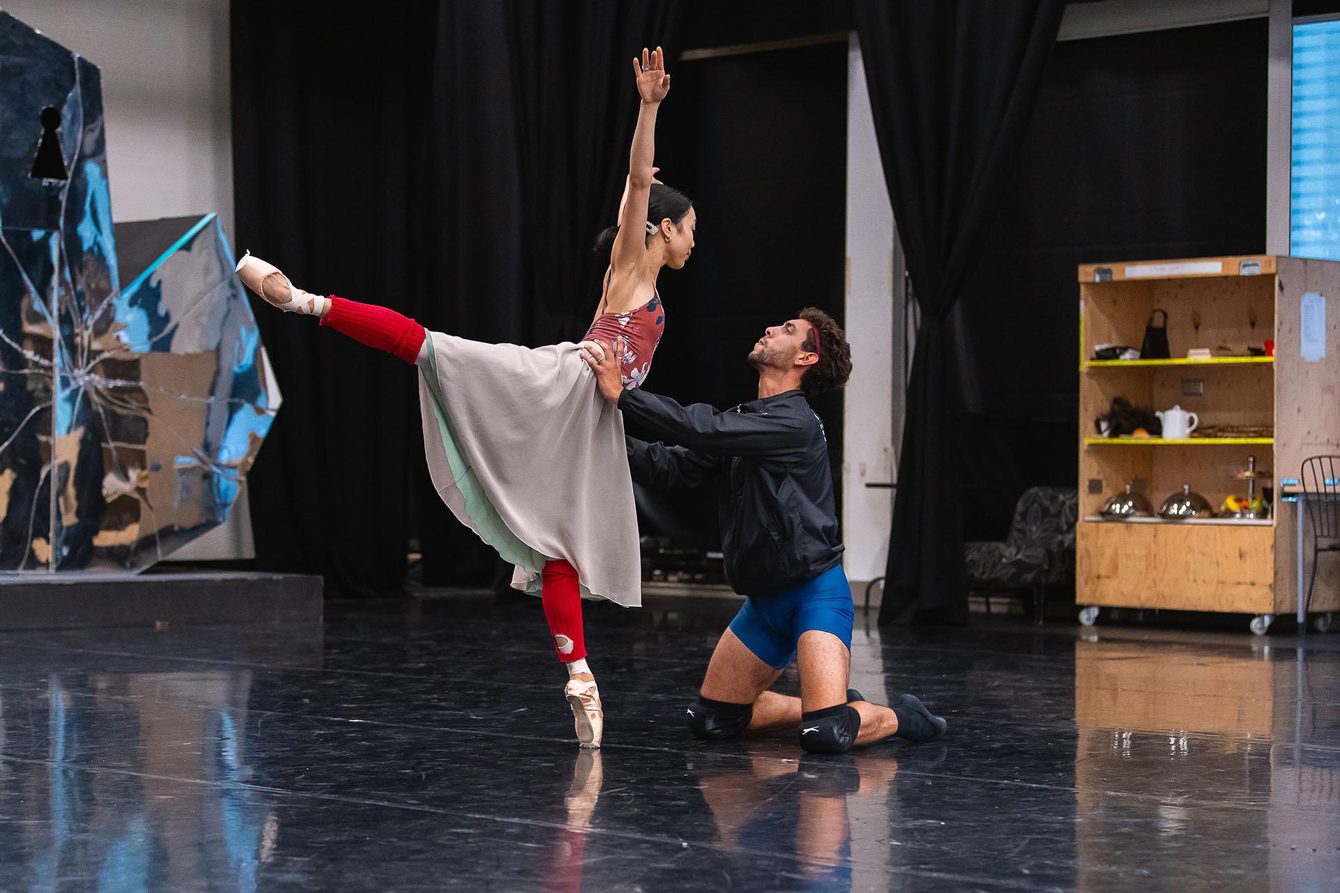 a female dancer stands on one foot en pointe, her leg perpendicular behind her and her arms raised high above her head. The male dancer, rehearsing as the Beast, kneels on the ground and holds her waist to support her.
