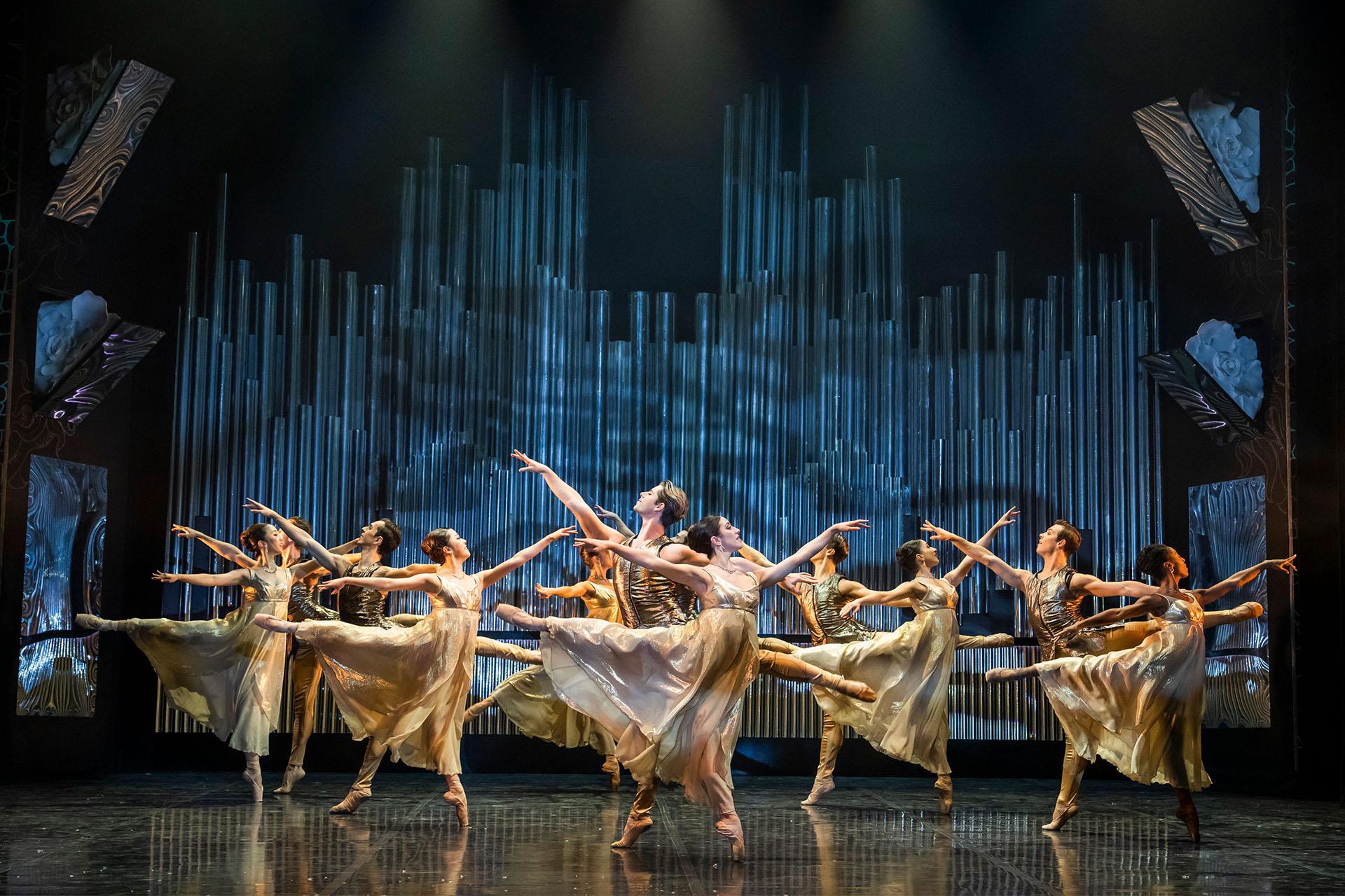 A dozen dancers in gold and white dance in unison, each stood on one foot, the other leg in arabesque behind them