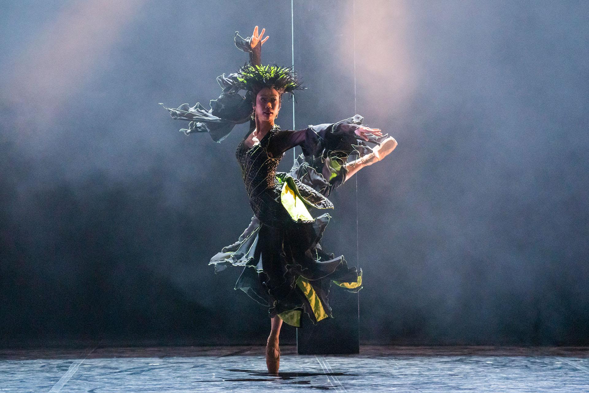 La Fée Magnifique standing on one foot en pointe, a leg raised behind her, one arm above her, her black and green dress spread out by the force of her spin.