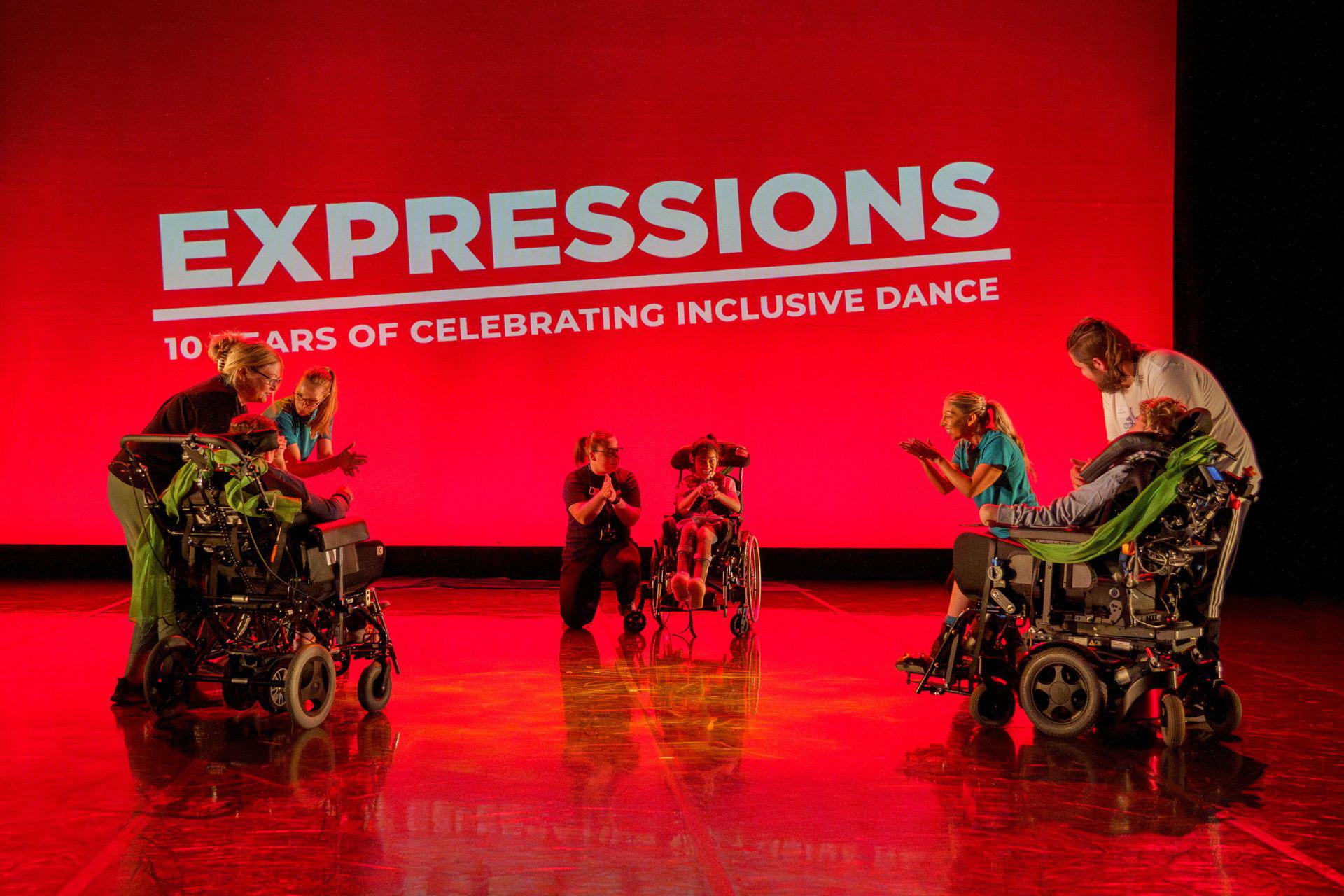 A group of children perform a wheelchair dance routine with their support workers on a red lit stage