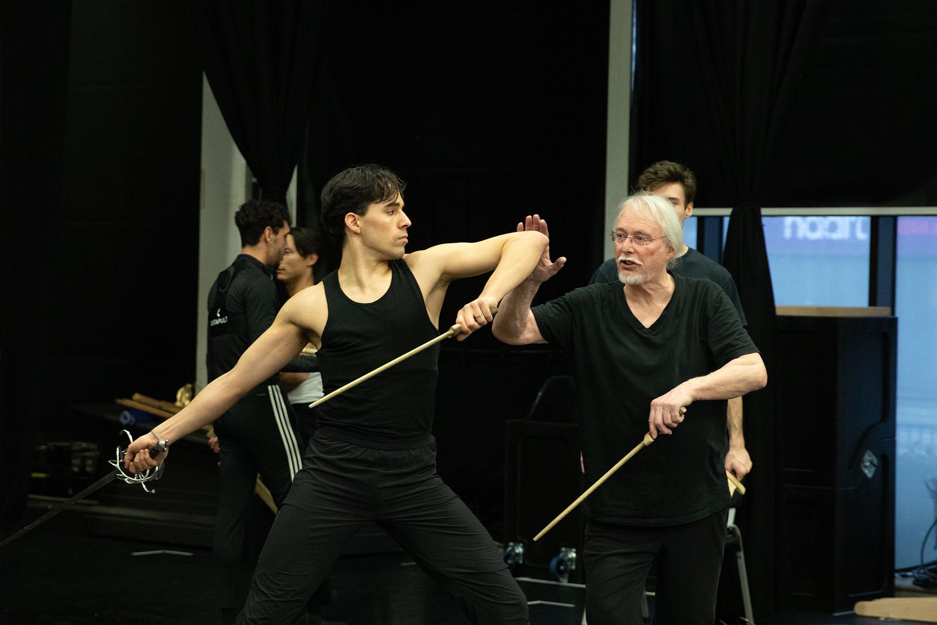 In rehearsals the grey haired fight director gives in struction to the dancer, placing his hand on his elbow instructively
