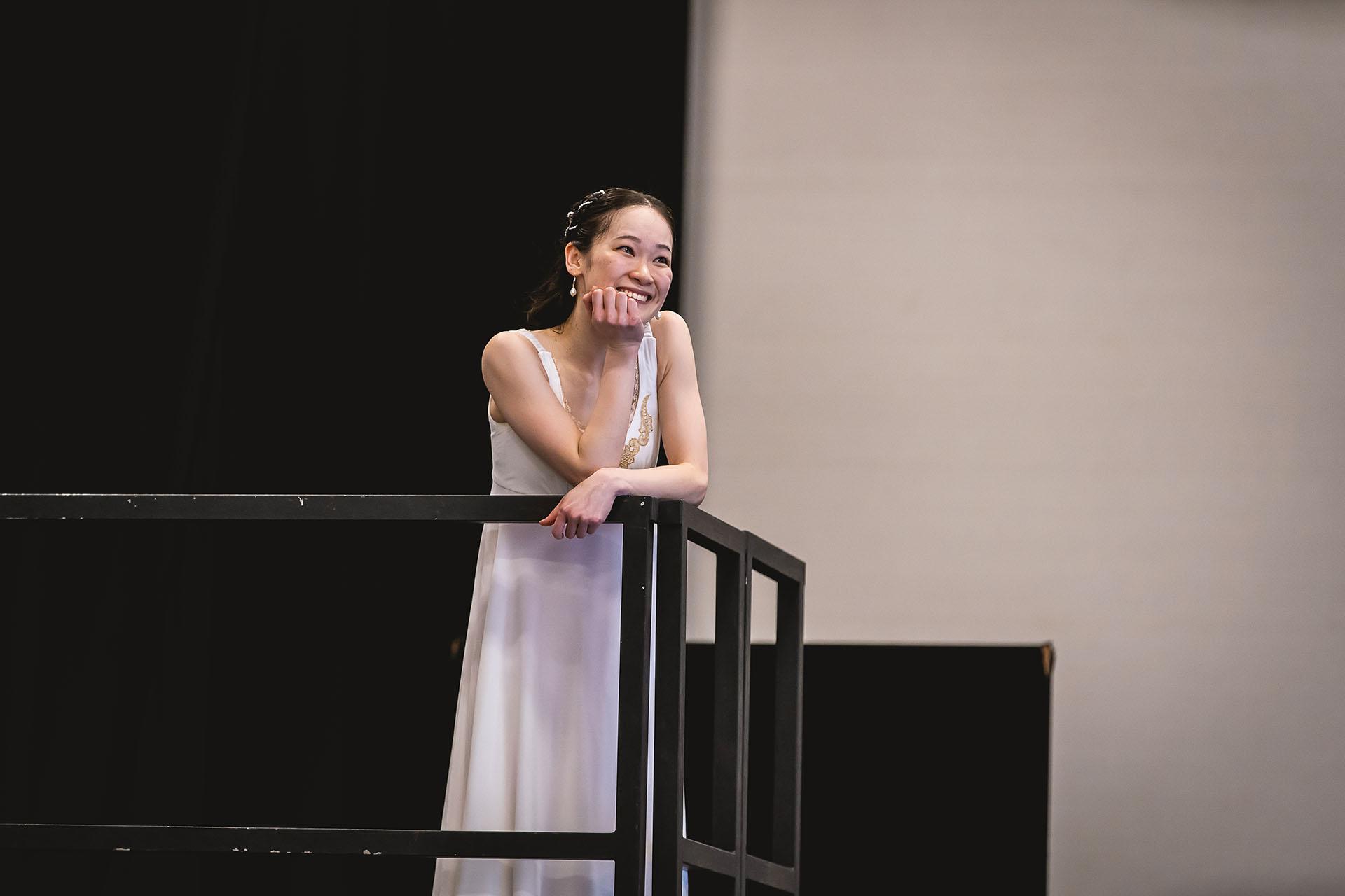 A dancer on a balcony dresses all in white smiles while resting a hand on her chin