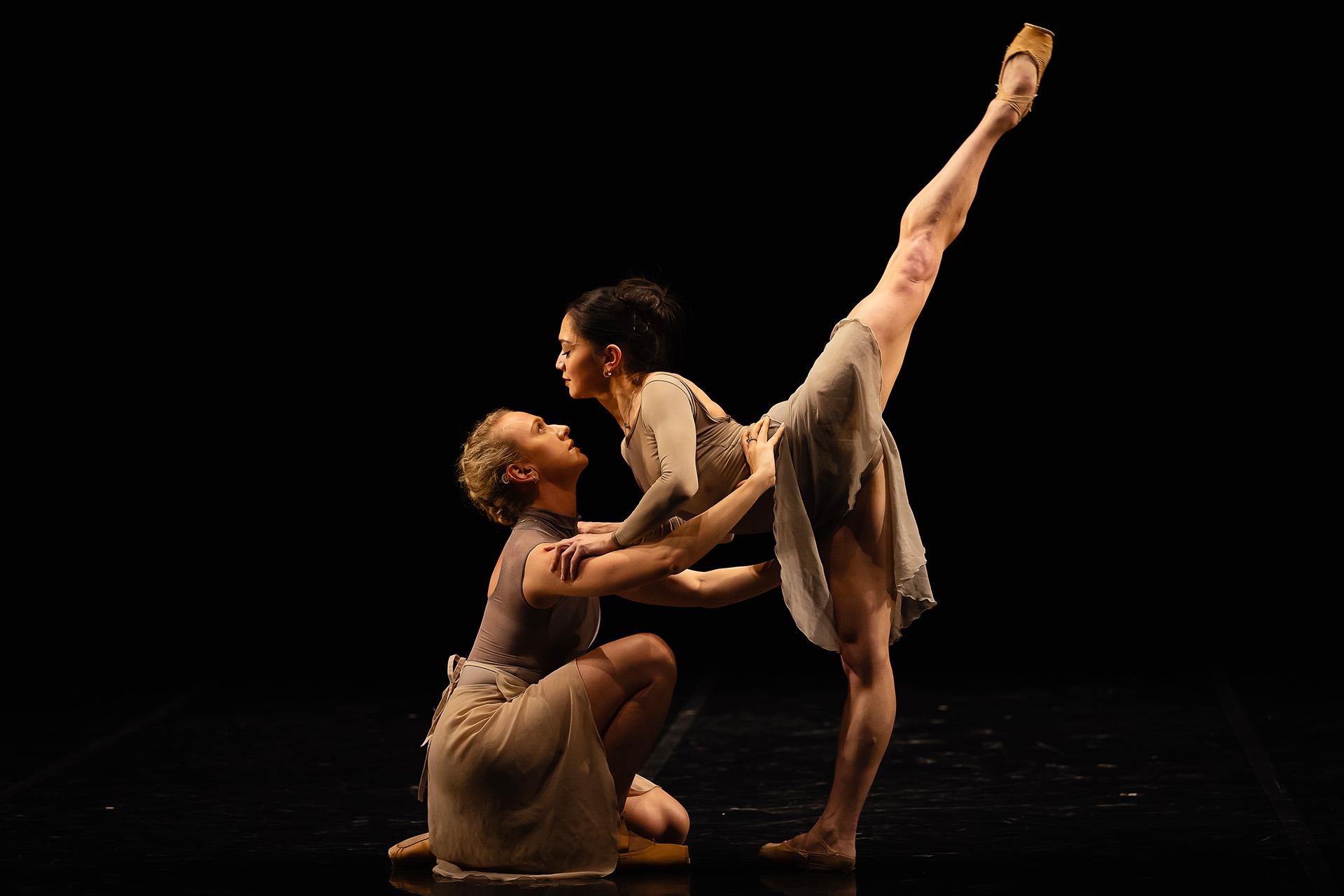 A dancer stands with one foot flat on the floow, the other leg raised straight up behind her as she leans her body forward and is supported by another dancer kneeling in front of her holding her waist