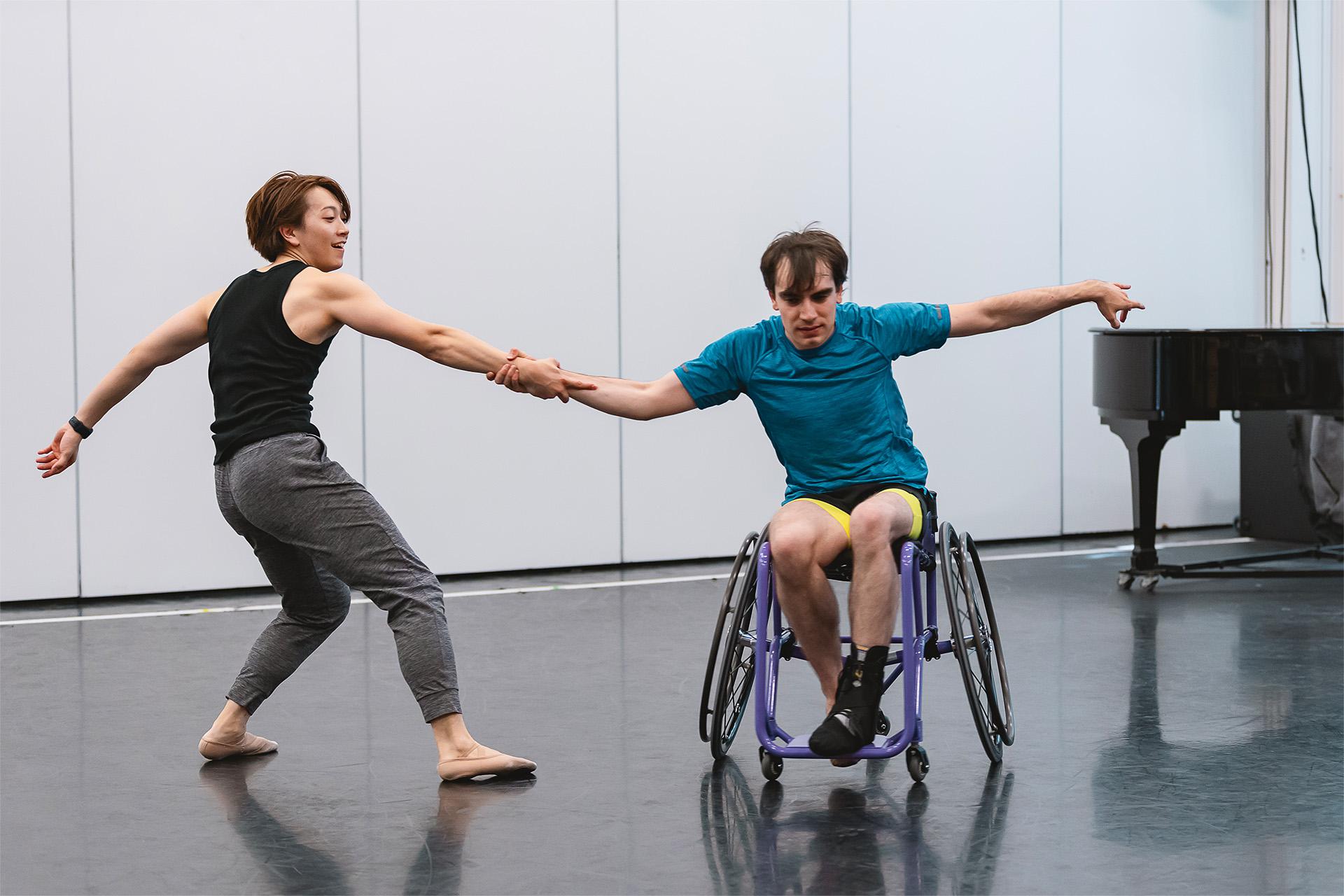 Two dancers hold hands and pull away from each other, one braced with his feet on the ground, the other with the wheels of his chair against the pull