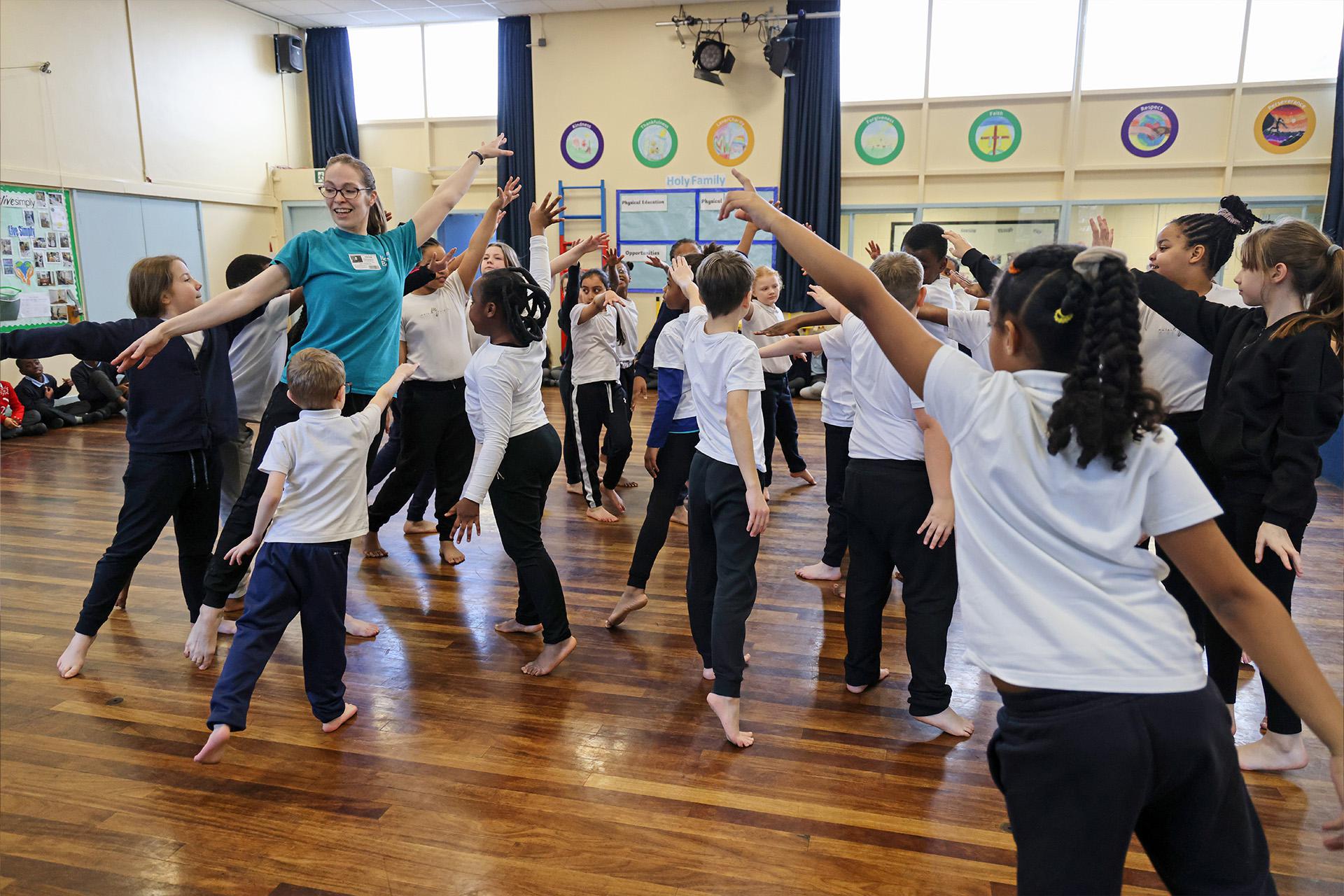 A group of children in a school hall, all barefoot, striking the same dance pose as each other - stretching one arm up and forward, the other down behind them as they step forward, their weight on the front foot