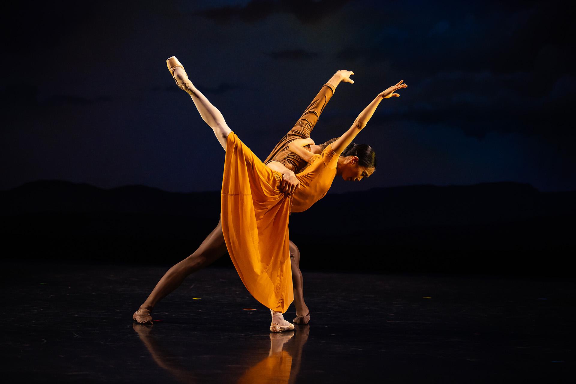 Two dancers, wearing umber and orange respectively, lean forward, one helping to balance the other as she stands arabeasque
