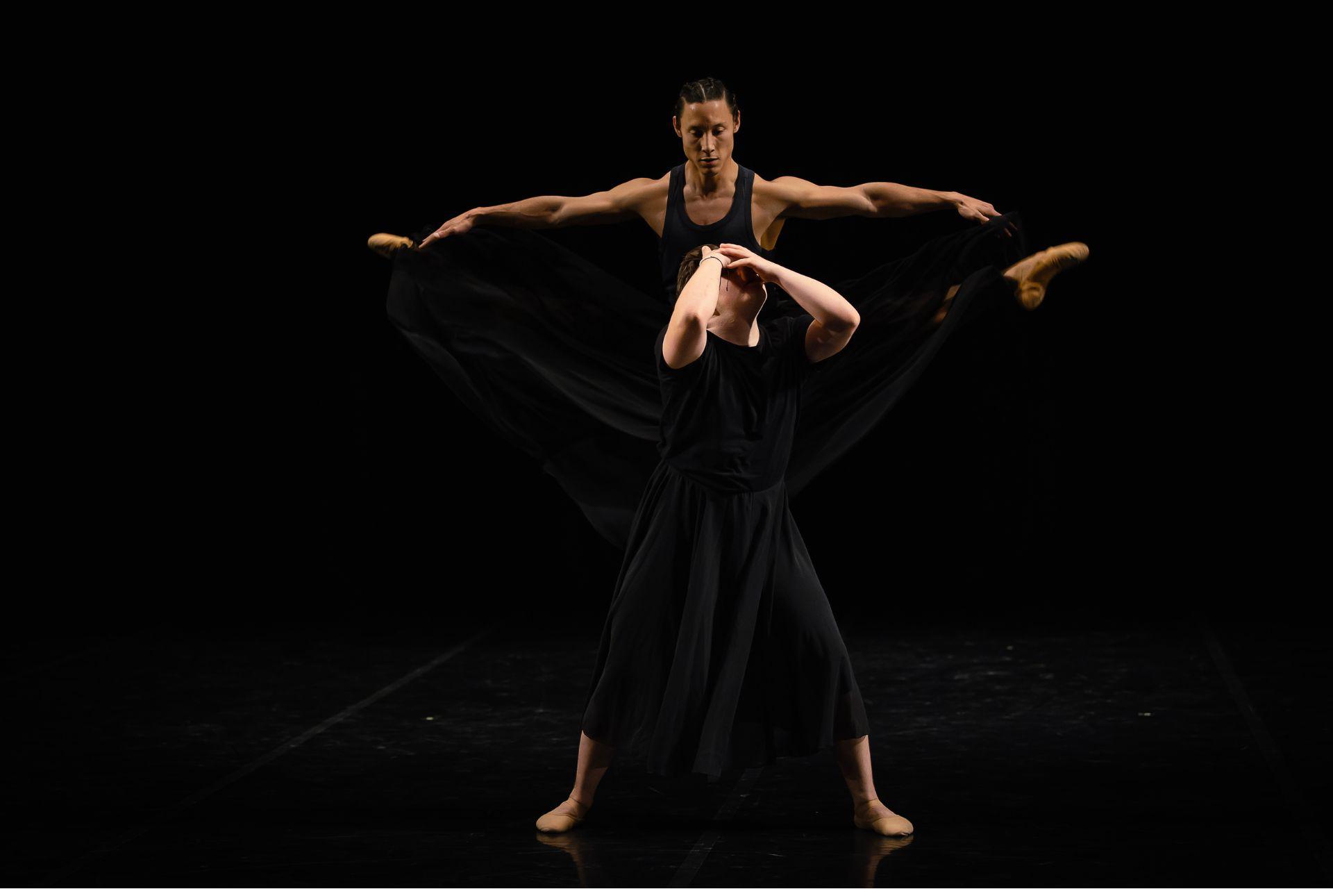 A dancer holds his face as another dancer leaps behind him with outstretched legs, looking down on the other dancer.