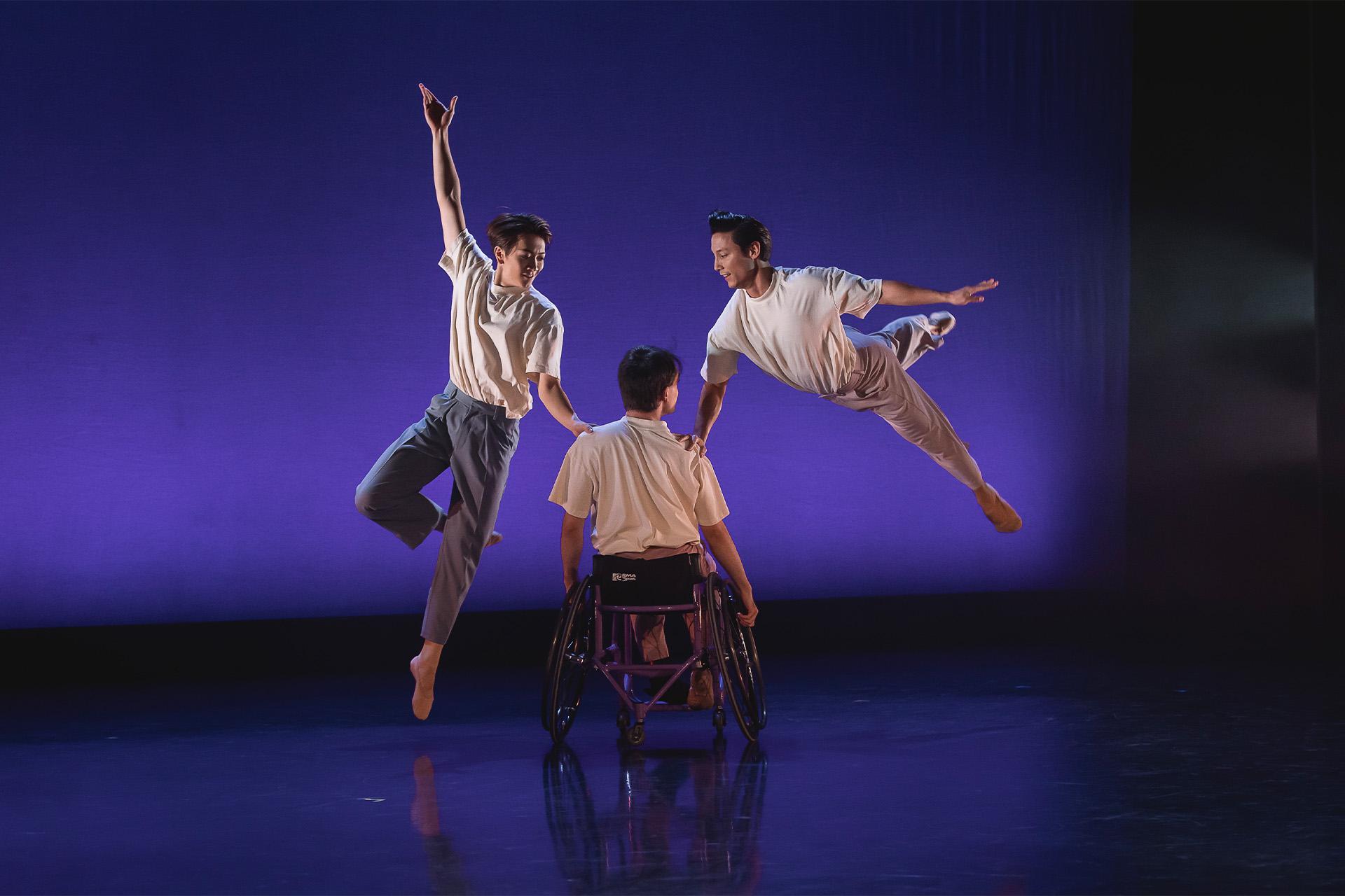 Three dancers on a purple stage, two dancers are leaping beside a dancer in a wheelchair
