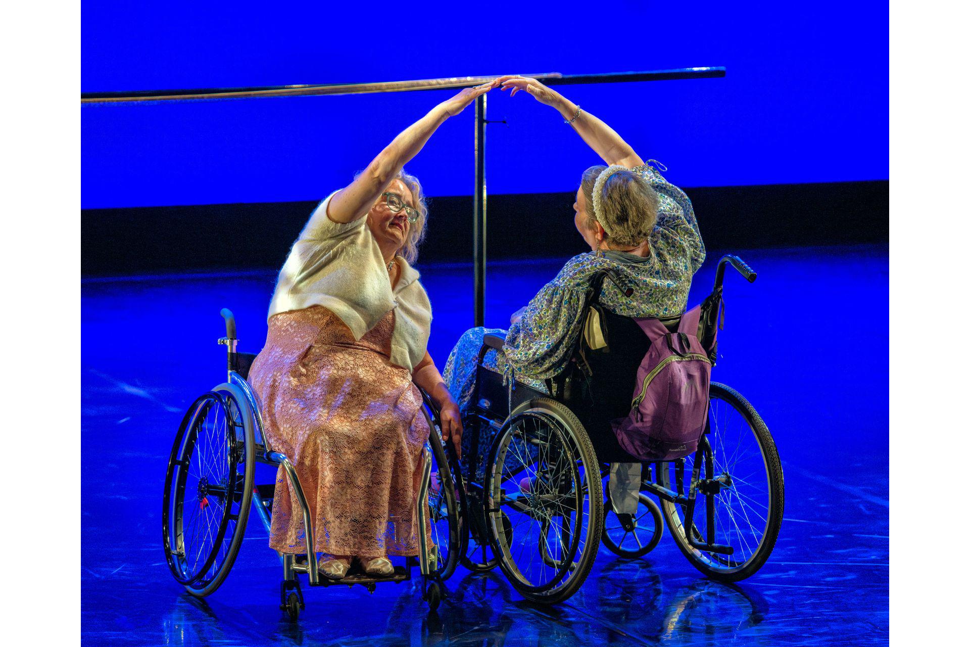 Two dancers in wheelchairs stand side by side, facing opposite directions. They lean towards each other raising an arm to touch fingers.