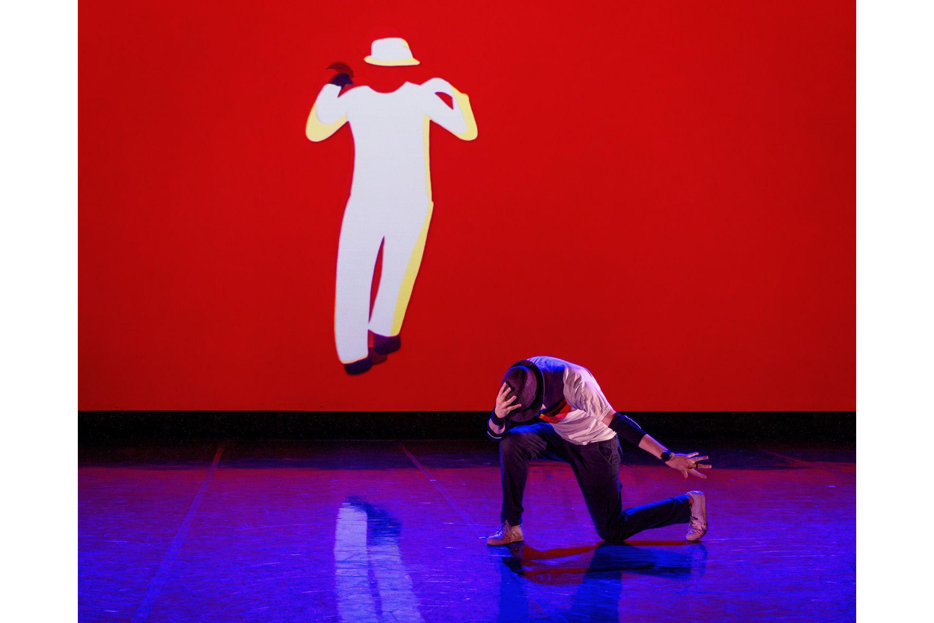 A dancer stands on one knee, leaning forward and holding his hat on his head. A white silhouette of himself can be seen on a red screen behind him