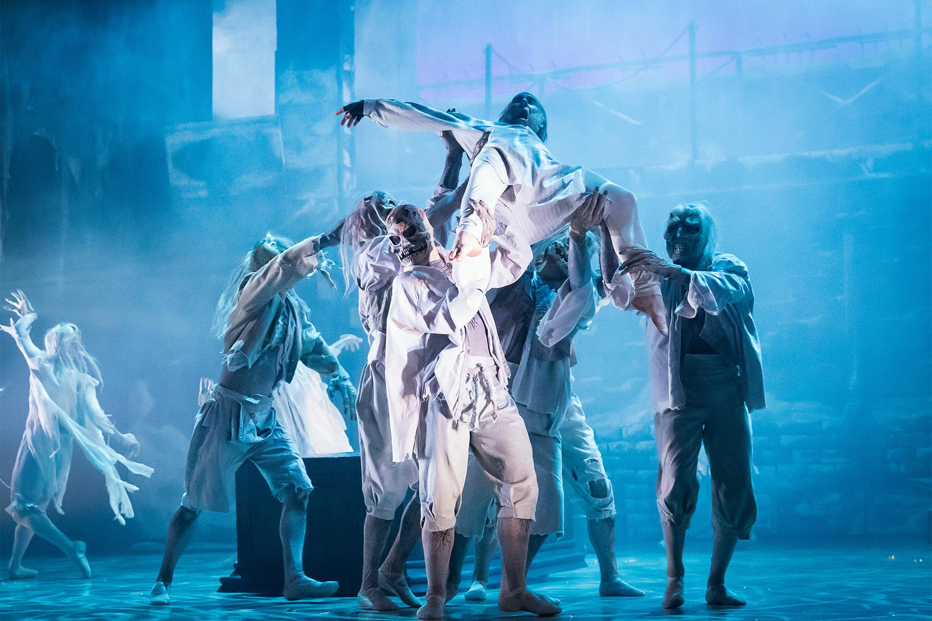 Dancers on stage dressed as phantoms lifting another dancer from a bed