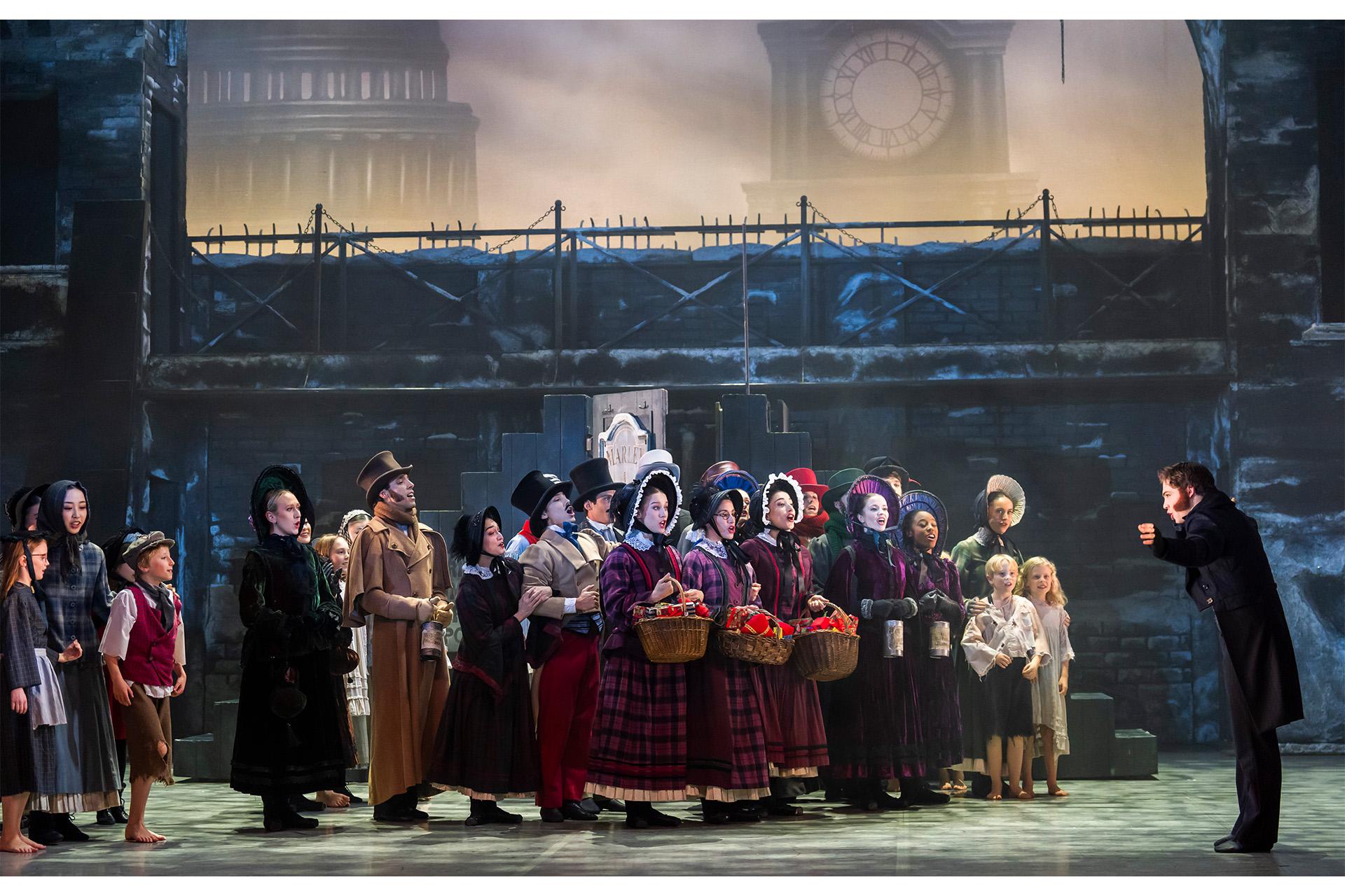 A large group of dancers in Dickensian costumes on stage singing