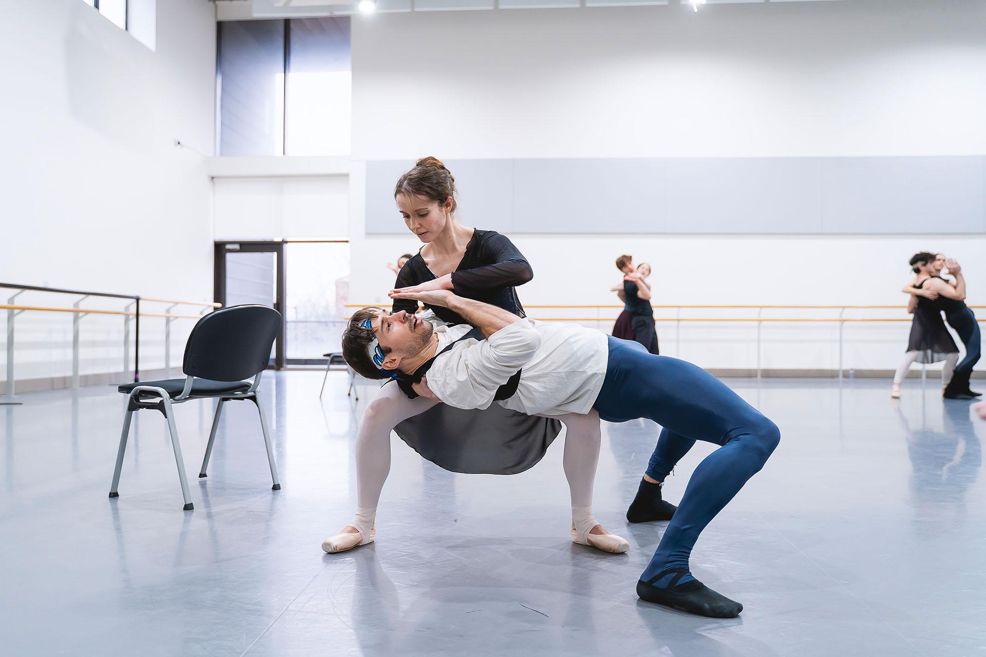 Male dancer leans back horizontally by his knees, supported by woman dancer with an arm under his shoulders, palms on their spare hand in from of his face touching