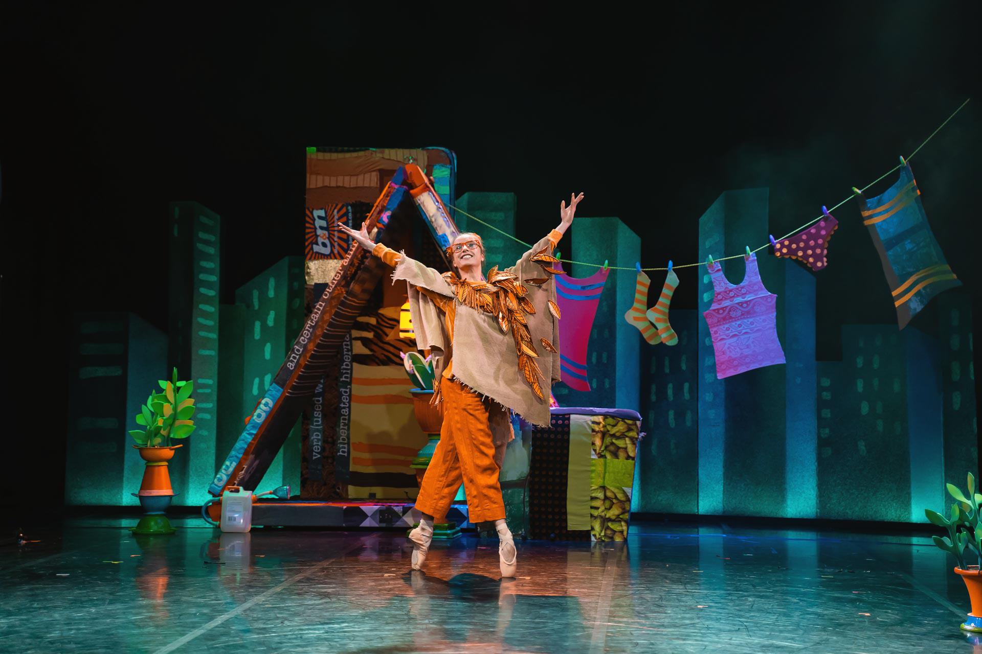 A dancer stood in orange clothes in front of a house with arms outstretched wide