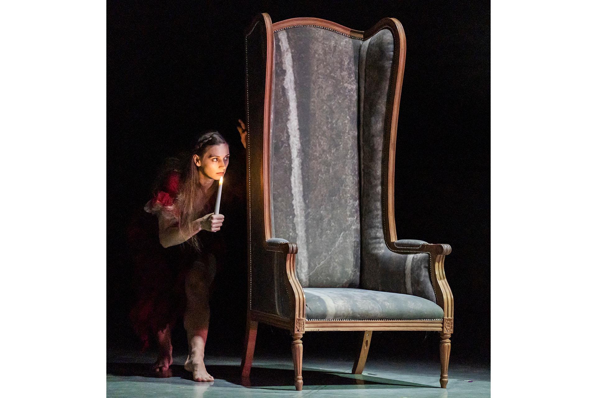 Lit by the flame of a candle, a mysterious figure, partly hidden by a large-backed chair