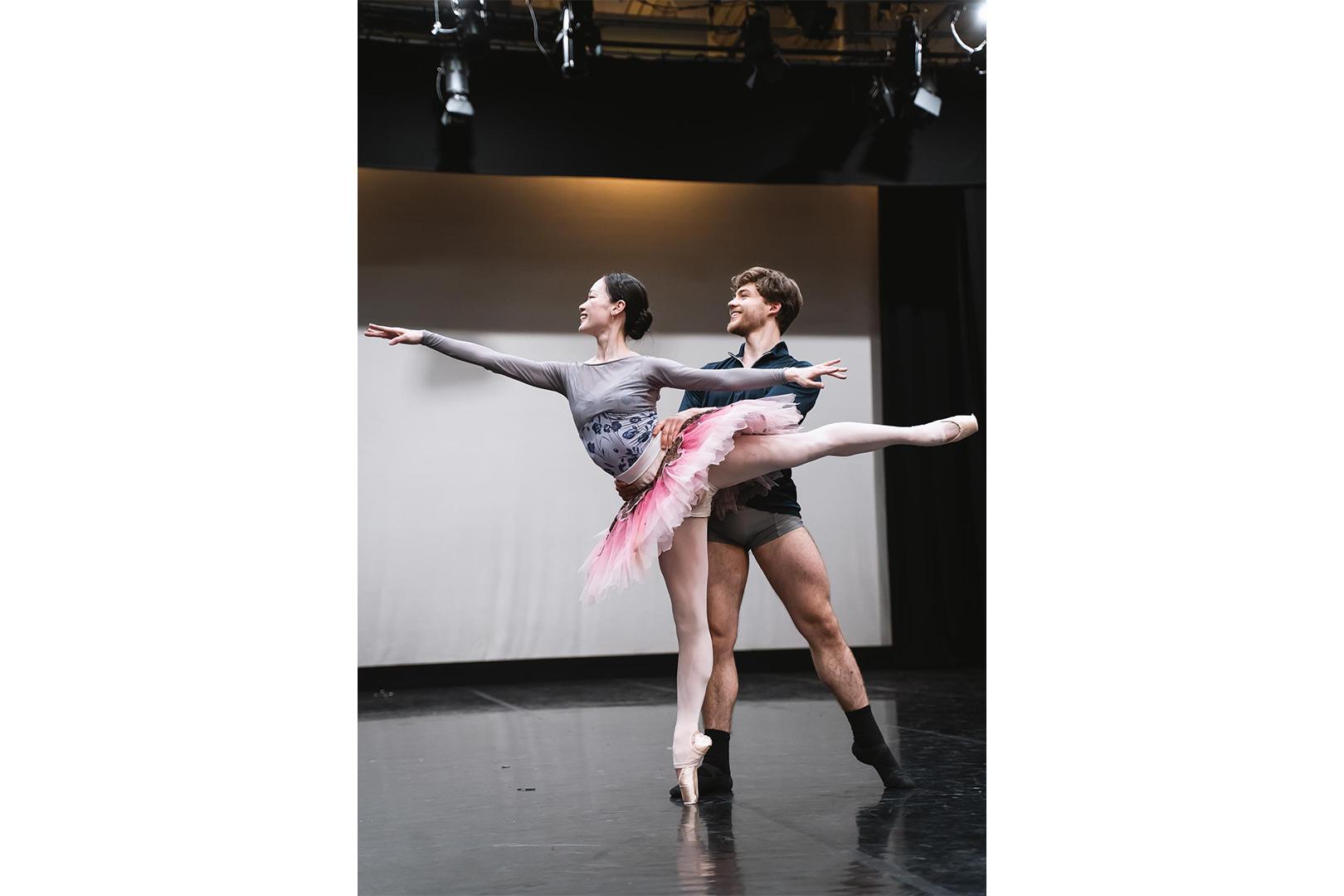 Two dancers, one in a pink tutu stood on pointe in an arabesque. The other dancer supports her