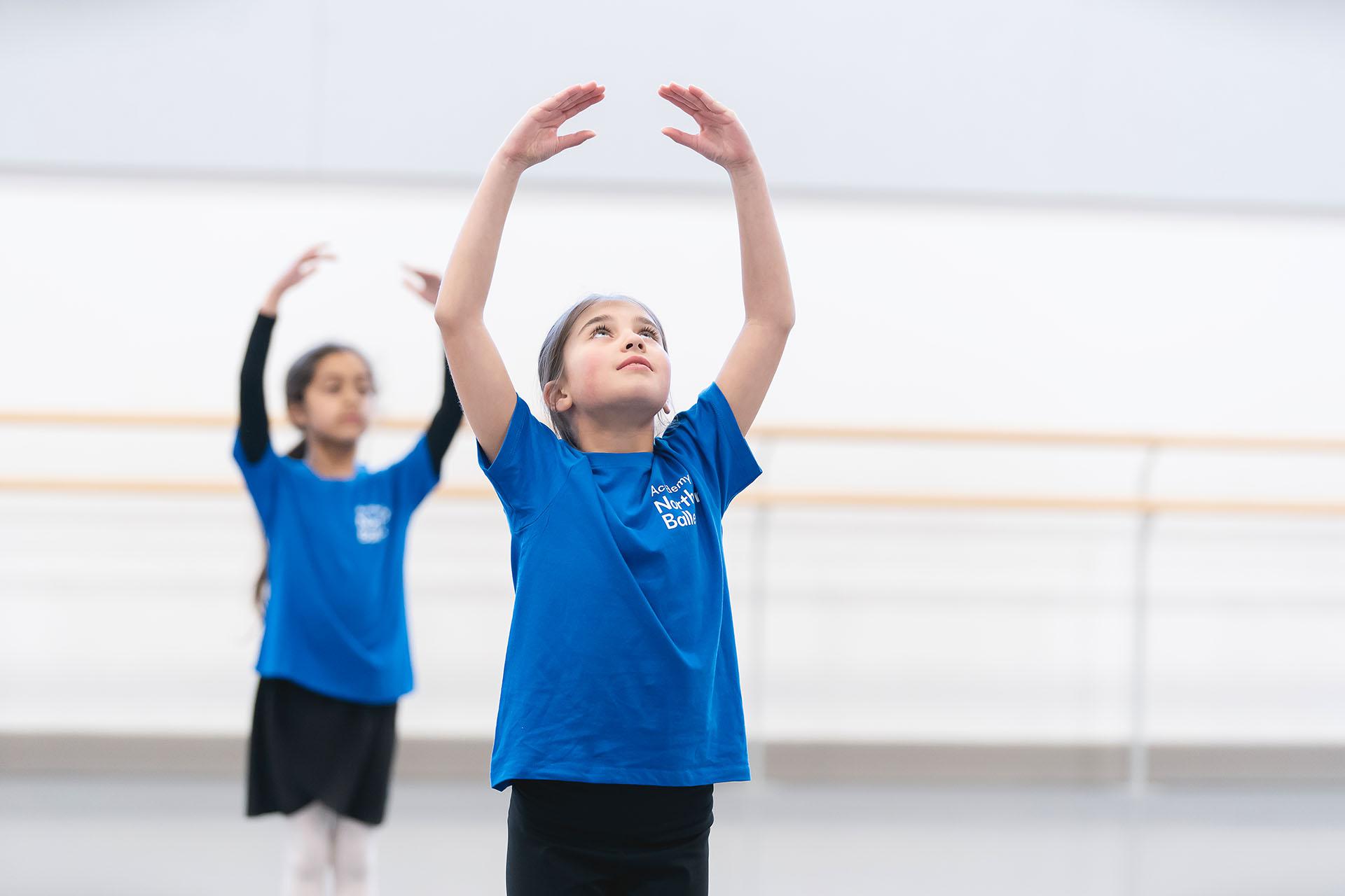 Child in a blue Academy of Northern Ballet T-Shirt stands with her hands over their head in fifth position