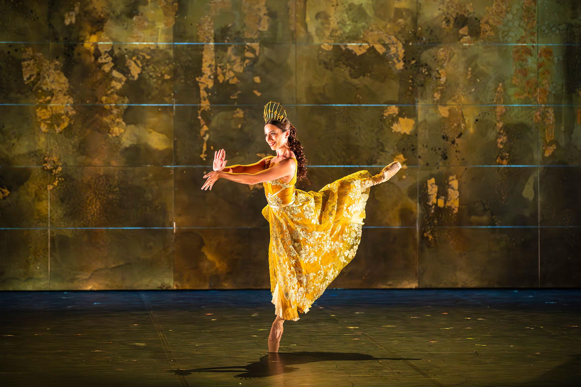Dancer in a yellow dress wearing a golden tiara stands arabesque
