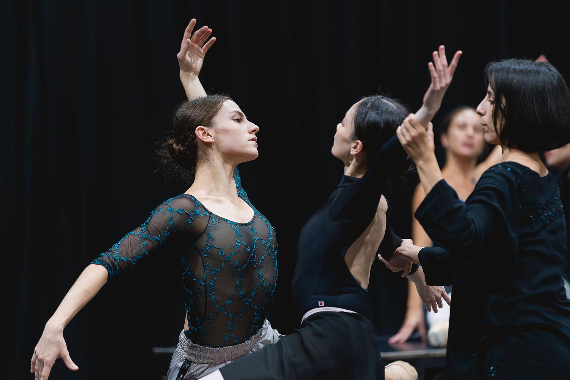 Two dancers face each other, mirroring moves, the choreographer behind one of them guiding her position