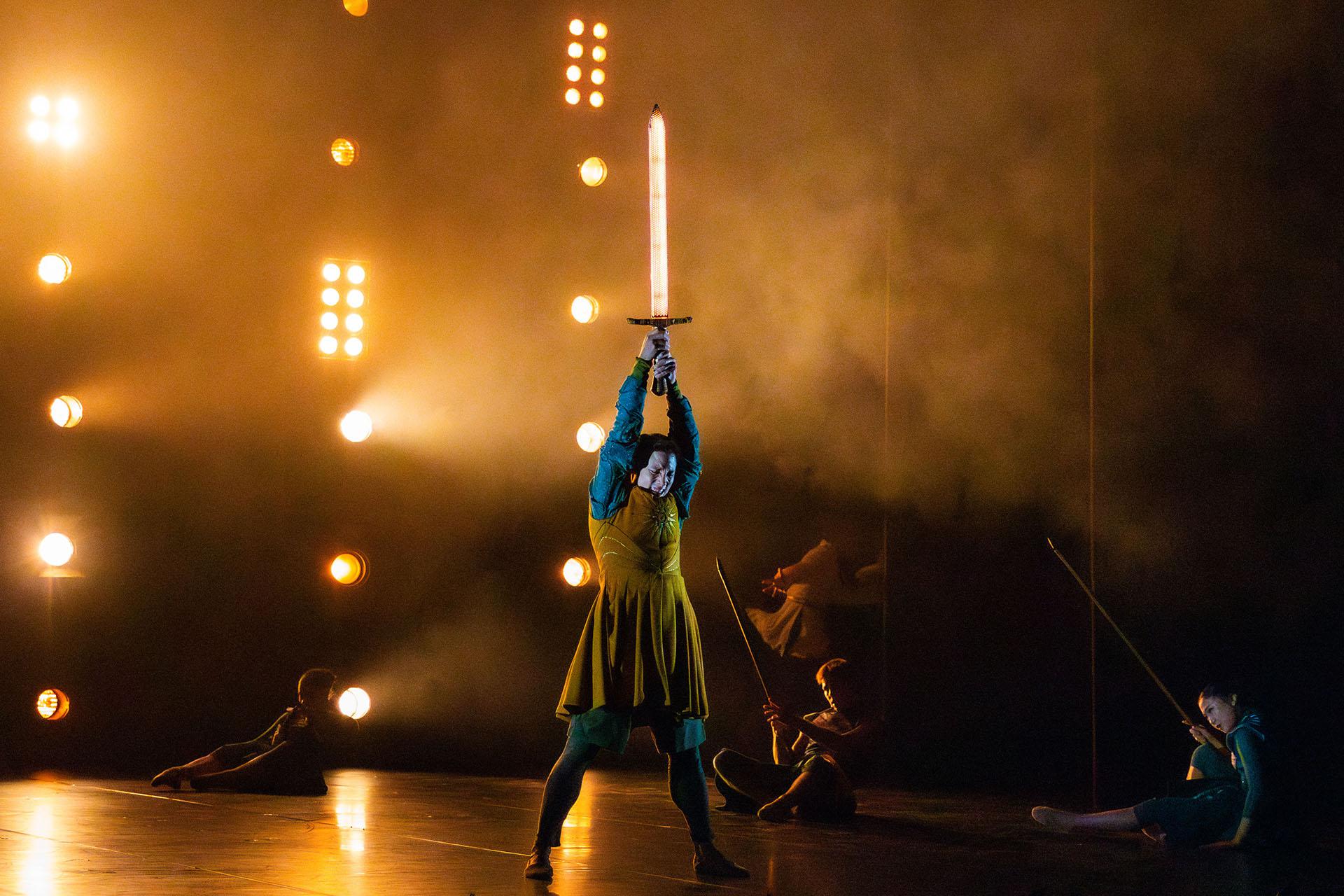 Wearing a yellow tunic with the symbol of the nation he is fighting with, Merlin holds a sword glowing with ethereal light above his head