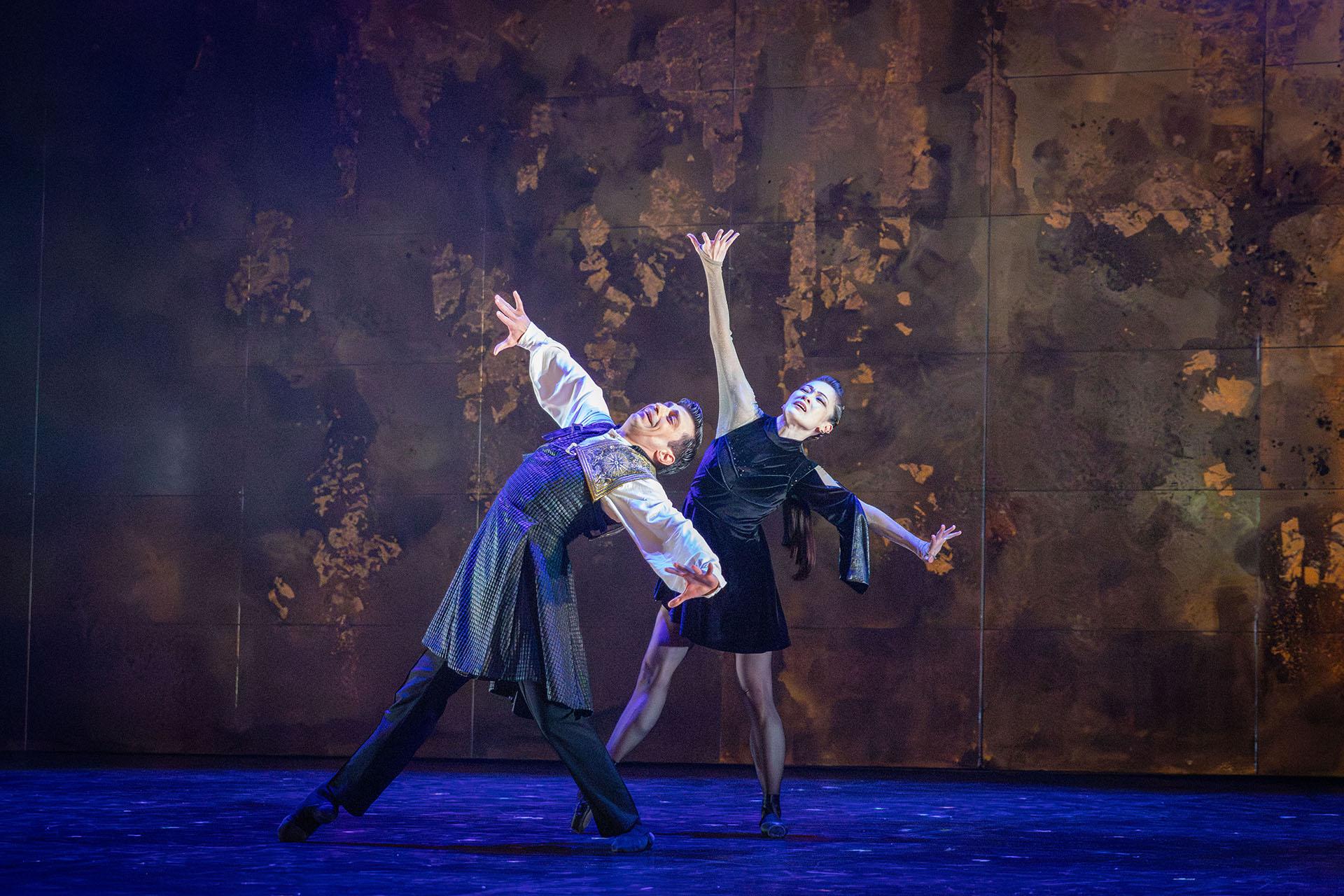 Two dancers dressed in black stand legs wide, leaning far back and their arms spread out from their body