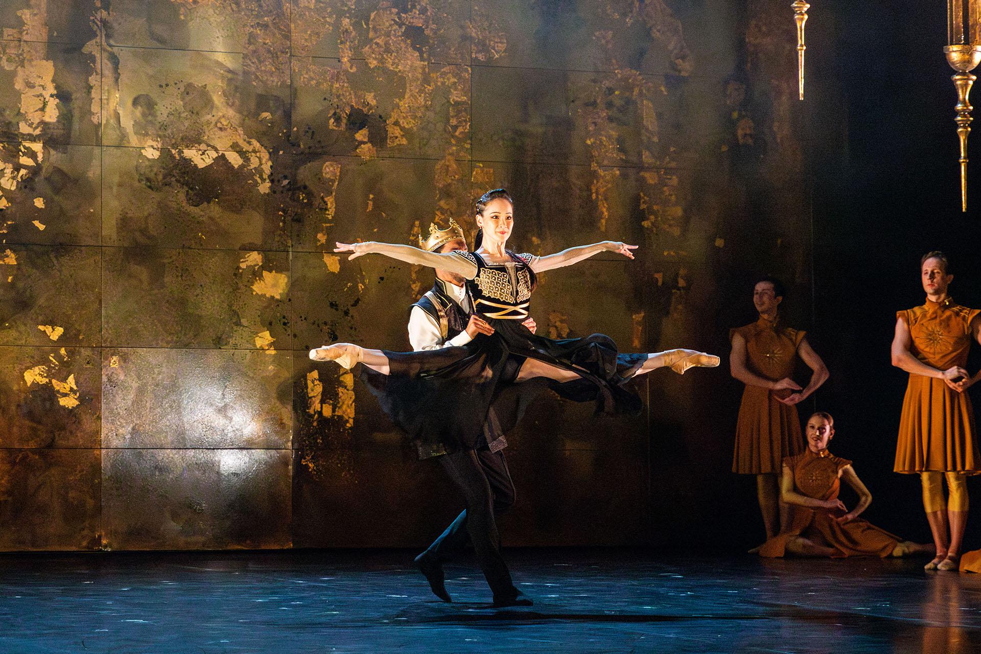 Morgan is lifted from the waist by a becrowned Vortigern, both dressed regally in black with gold brockade.