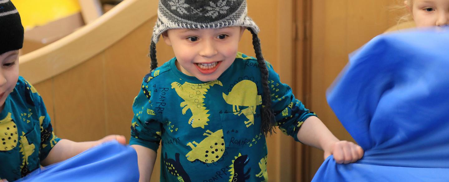A young child smiling wearing a wooly hat and a green jumper with yellow and black dinosaurs