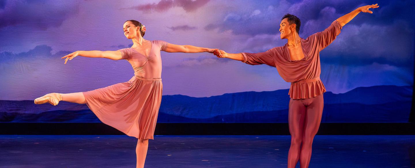 A male and female dancer hold hands. The female dancer lifts her leg and reaches to the left, as the male dancer floats his arm upwards and smiles at her