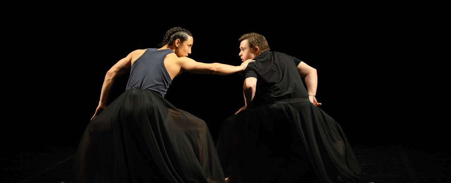 Two dancers stand hands on their bent knees. They look at each other as one grabs the other's shoulder.