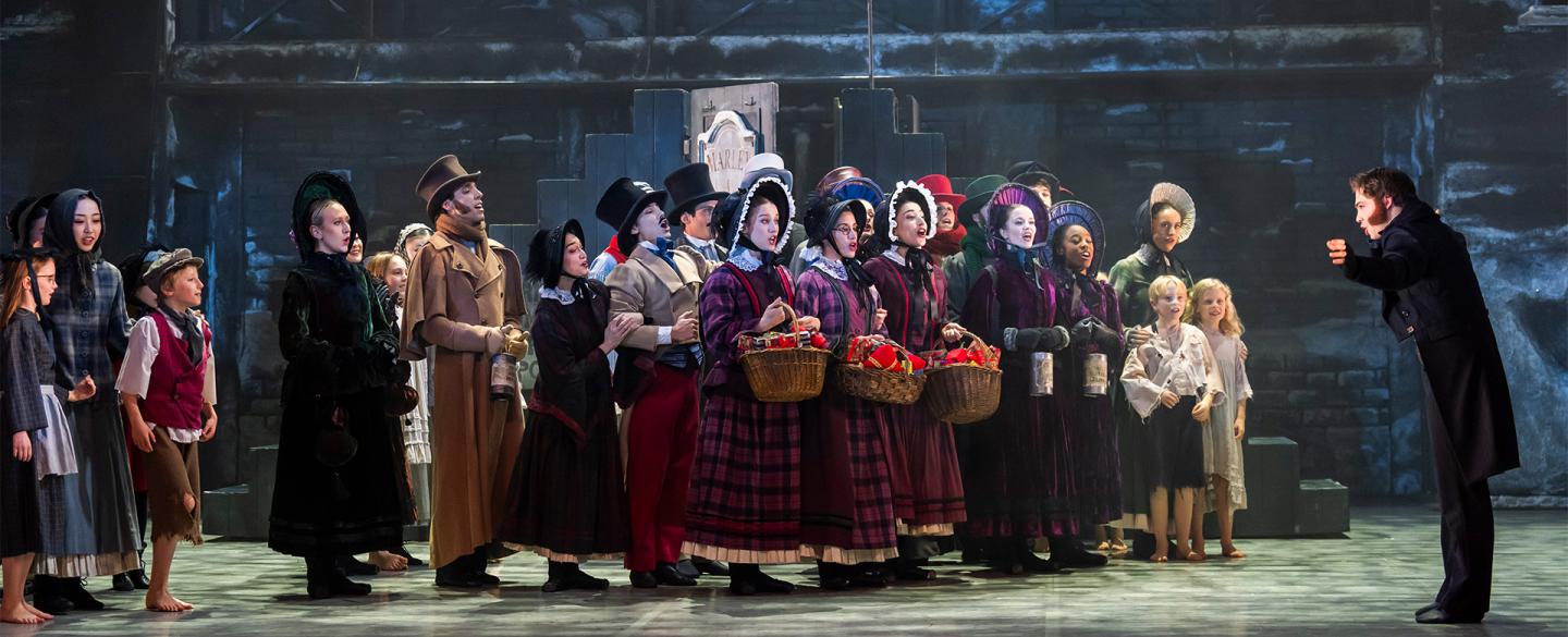 A large group of dancers in Dickensian costumes on stage singing
