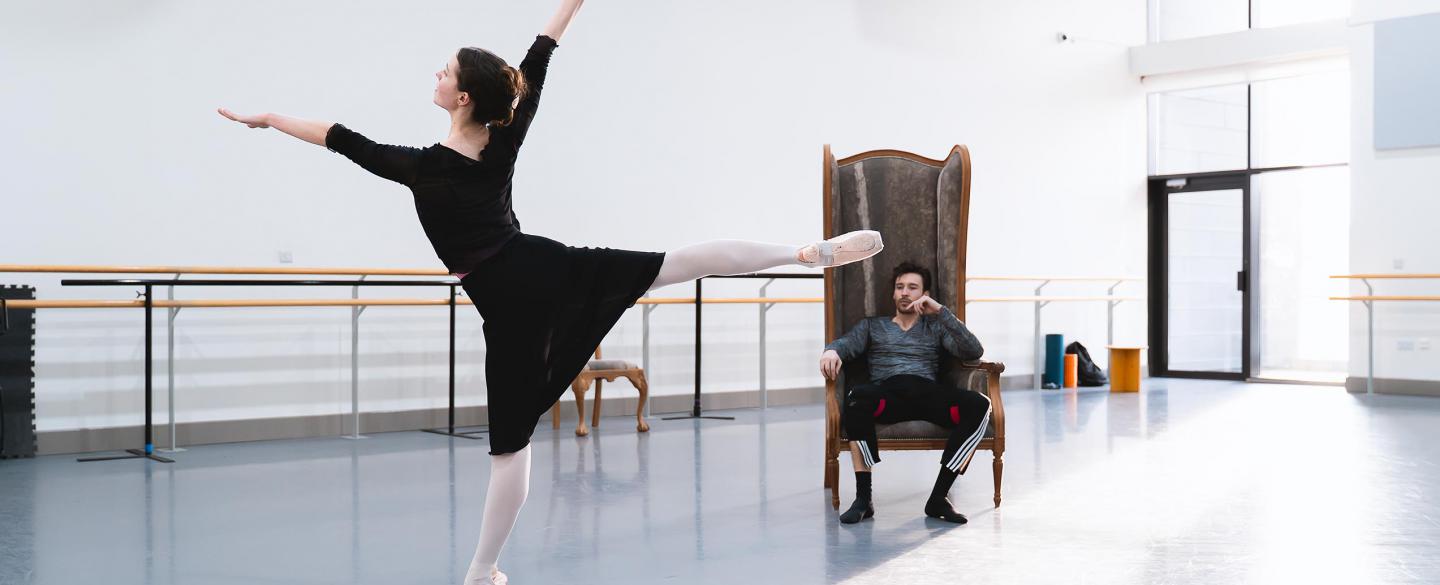 A dancer stood on pointe with legs and arms outstretched with another dancer watching on in a large armchair