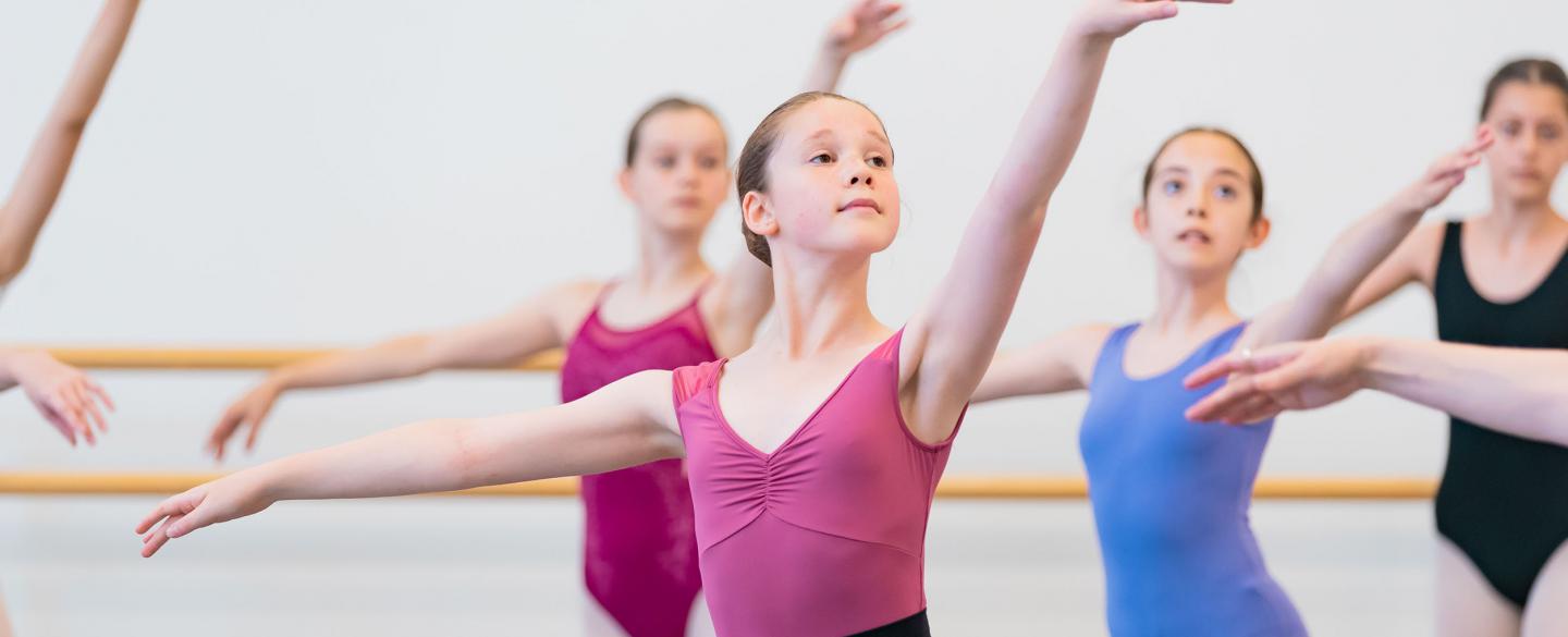 A young girl in a pink leotard stands in a ballet class with arms outstretched