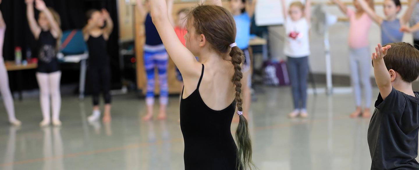 Girl in black leotard stands in a dance studio with her arms raised, in the background more children stand with their arms raised