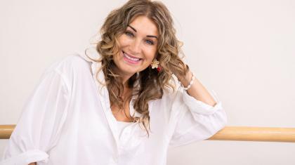 Woman in white blouse, long curly light-brown hair, relaxing against a ballet barre smiling