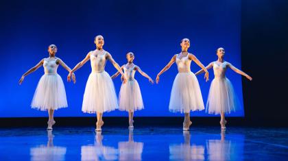 Students in white perform on a stage, standing on their toes with their arms at their side.