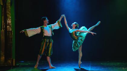 Two dancers stood in a traditional ballet pose. One dancer is on pointe with leg raised high