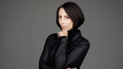 Woman with dark hair, a wan smile, black jacket and her arms crossed with one hand under her chin